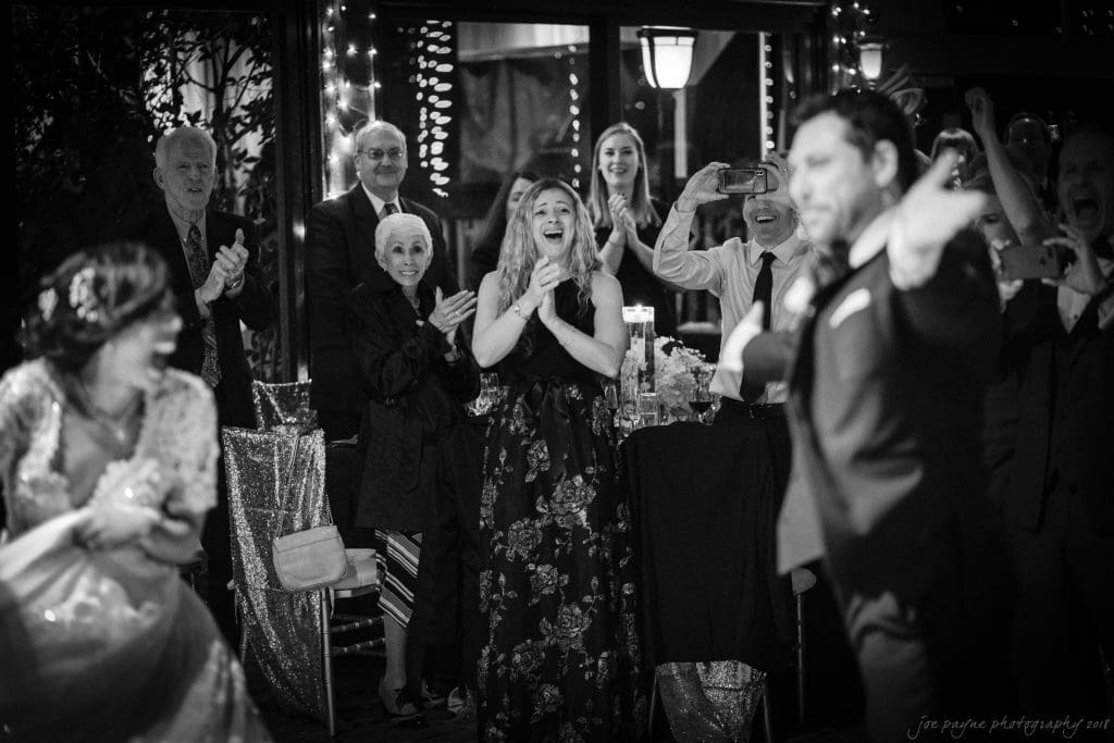 biltmore wedding photographer - anneké & andrew A black and white photo of a wedding reception. Guests stand and clap joyfully as a bride and groom dance. The room is decorated with lights and a floral arrangement. People are smiling and capturing the moment with phones and cameras.