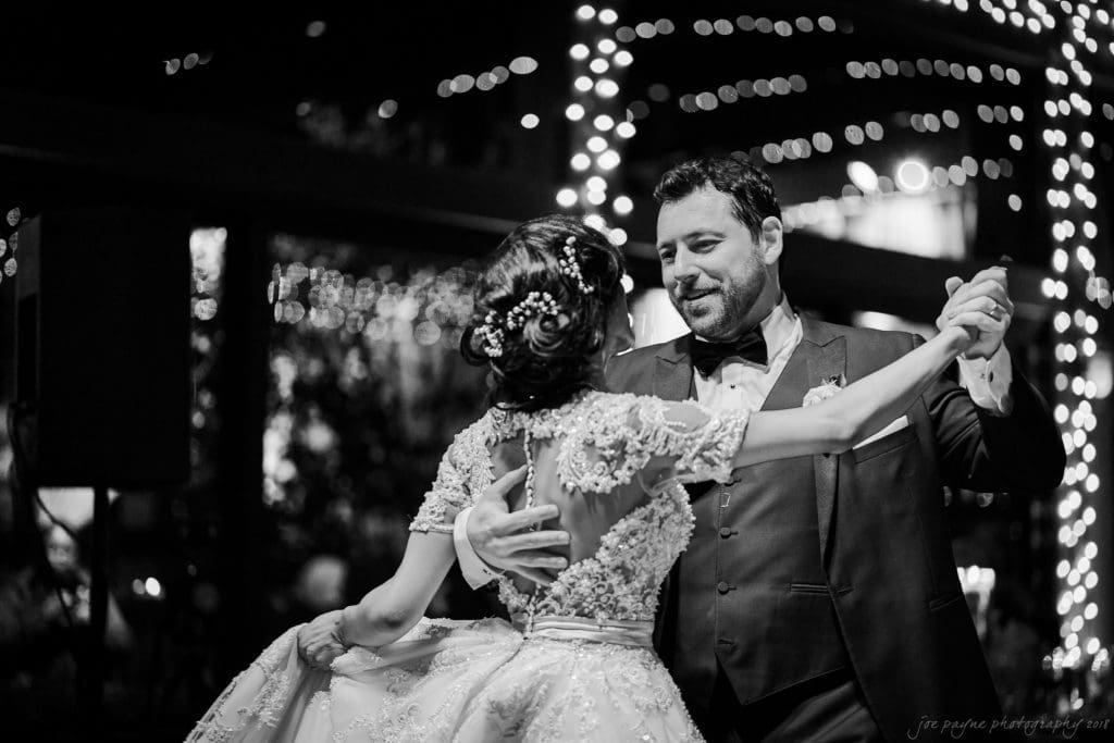biltmore wedding photographer - anneké & andrew A bride and groom dance together under twinkling lights at night. The groom wears a suit and bow tie, while the bride is in an elegant gown with floral details. The setting is festive and romantic.