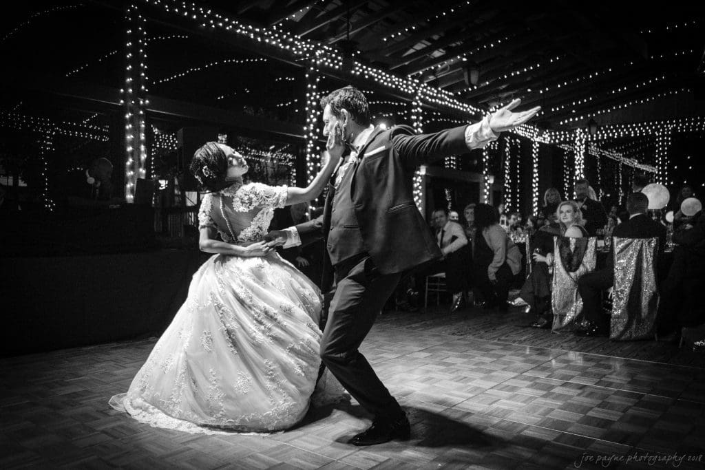 biltmore wedding photographer - anneké & andrew A bride and groom gracefully dance under string lights in a dimly lit venue. The bride wears an elegant gown, and the groom is in a suit. Guests seated around watch the performance, adding to the romantic atmosphere.
