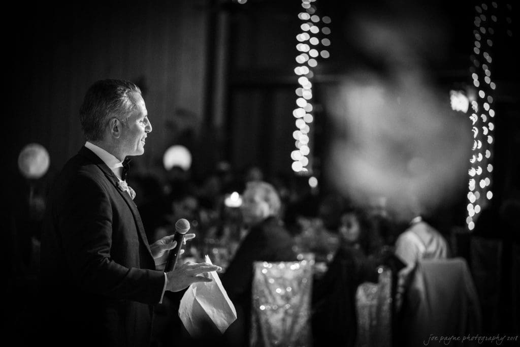 biltmore wedding photographer - anneké & andrew A man in a suit holds a microphone while speaking at an event. The setting is dimly lit with festive lights in the background, and people are seated at tables, listening attentively.