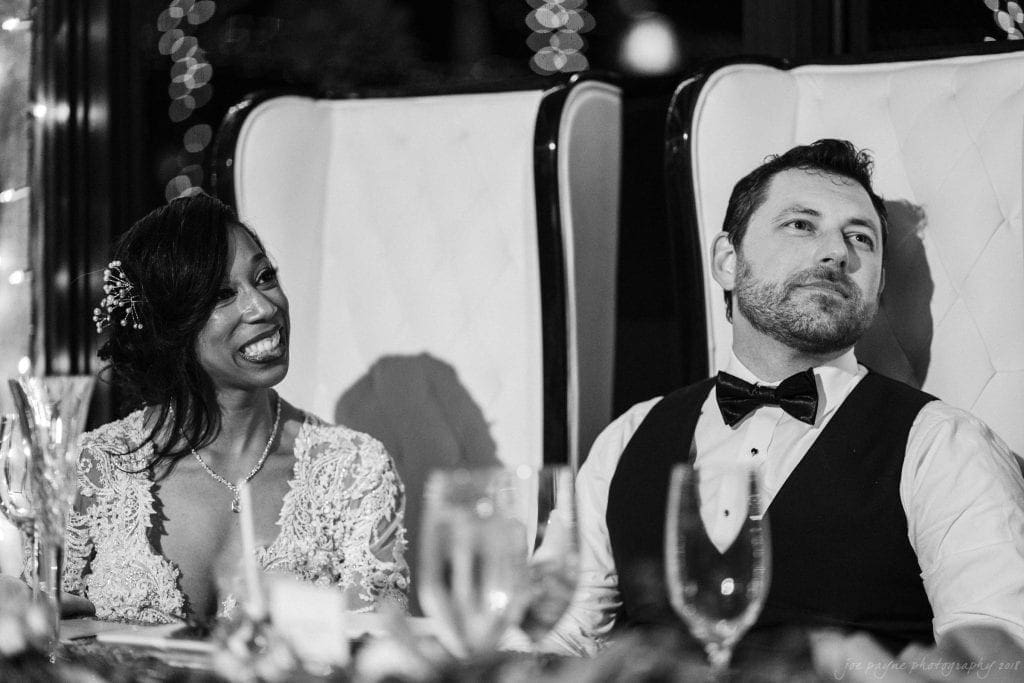 biltmore wedding photographer - anneké & andrew A black and white photo of a happy couple sitting together. The woman is wearing a lace dress and smiling, while the man, dressed in a vest and bow tie, looks content. They are seated in high-backed chairs at a formal event.