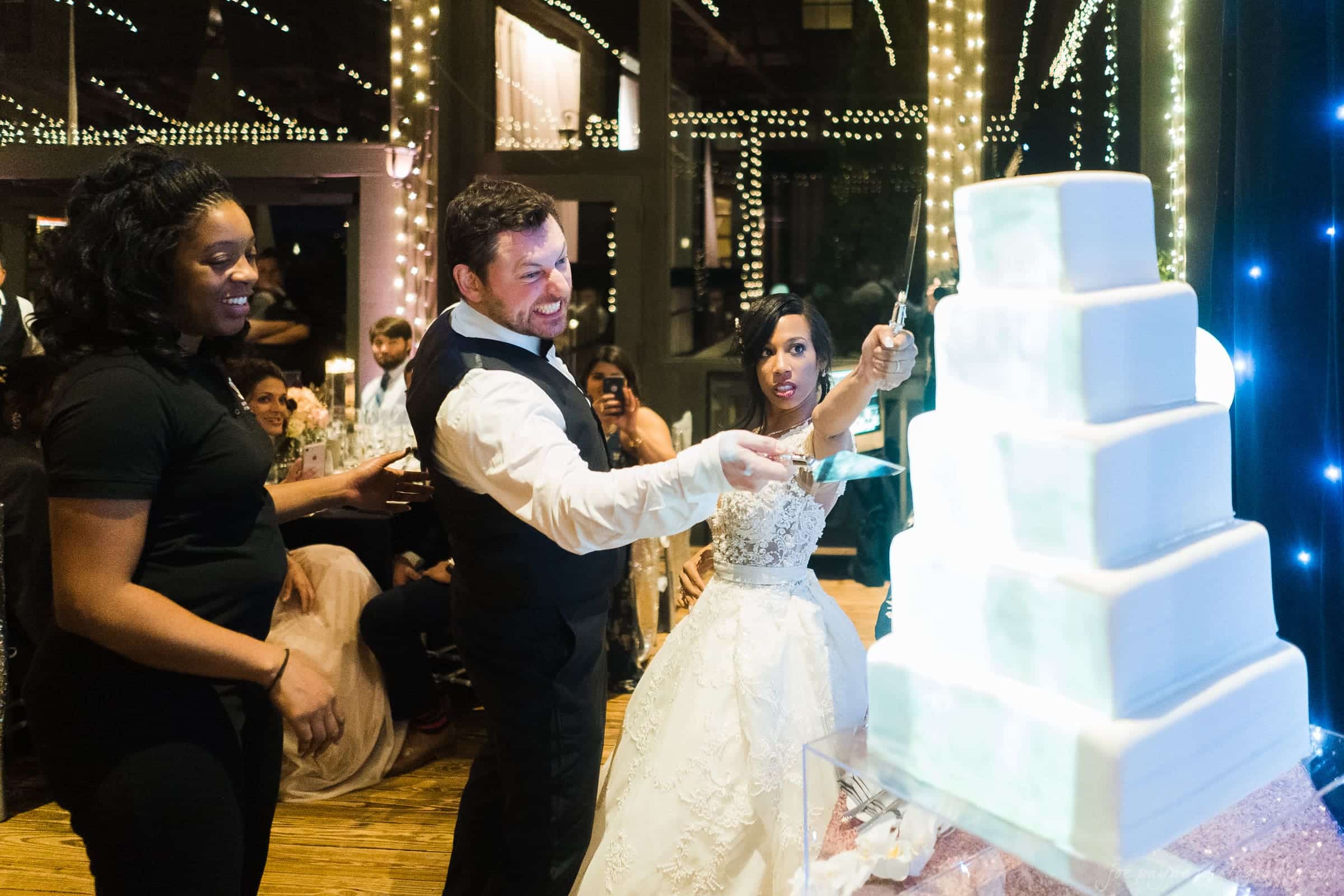 biltmore wedding photographer - anneké & andrew A bride and groom are cutting a large, tiered white wedding cake together. They are surrounded by guests at a warmly lit venue, with string lights in the background. The bride holds the knife, smiling excitedly, as the groom guides her hand.