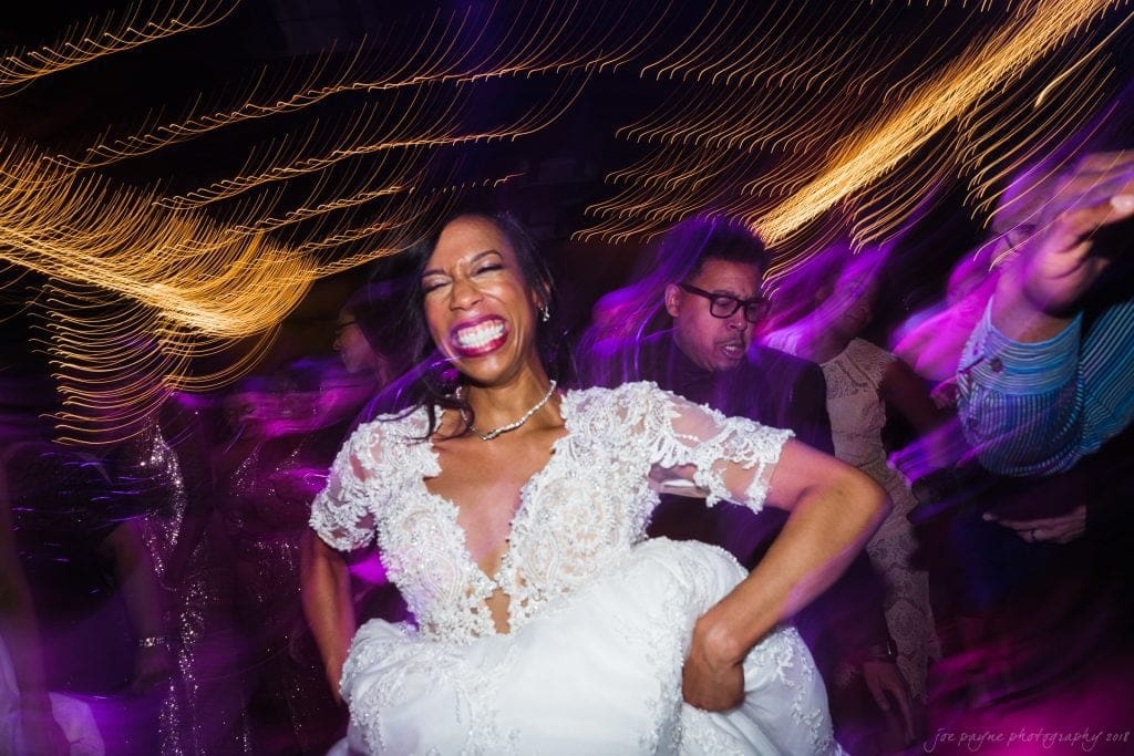 biltmore wedding photographer - anneké & andrew A joyful woman in a white dress is dancing energetically in a lively party setting, surrounded by vibrant swirling lights and dark purple hues, creating a dynamic and festive atmosphere.