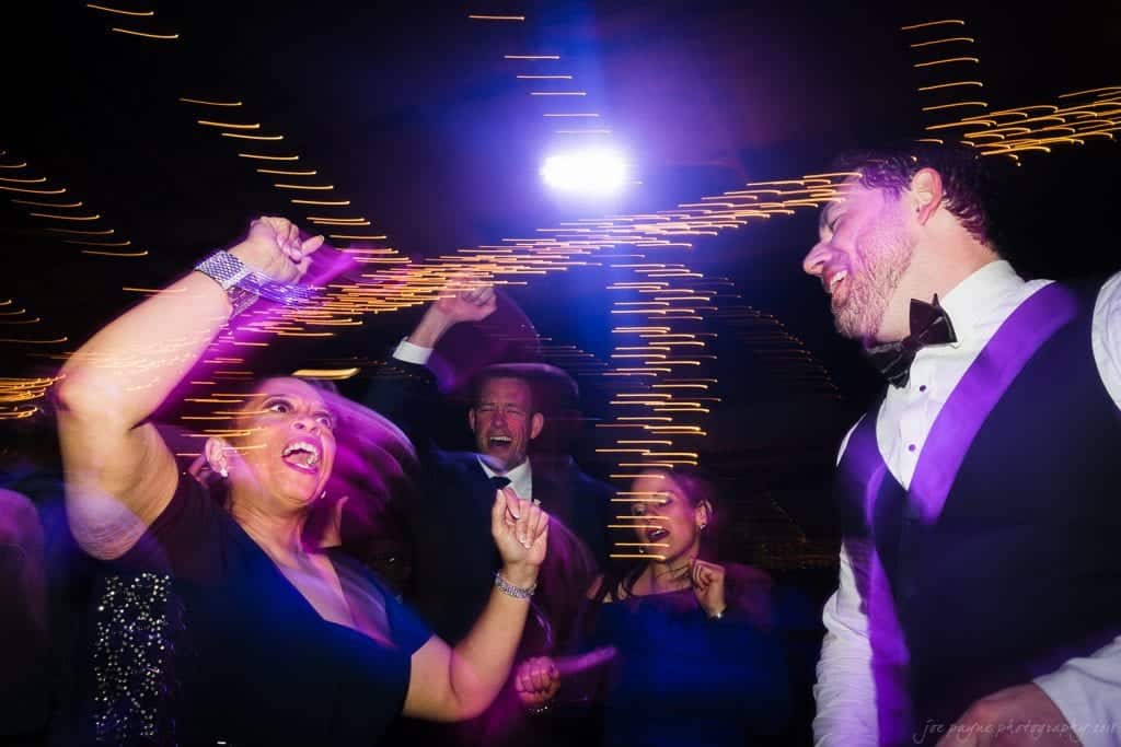 biltmore wedding photographer - anneké & andrew People dancing energetically at a party with colorful, blurred lights creating a dynamic atmosphere. The scene captures joy and movement, with a man in a suit and others in festive attire enjoying the lively event.