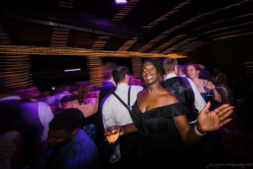 biltmore wedding photographer - anneké & andrew A joyful woman in a black dress dances with a drink in hand, surrounded by a lively crowd at a party. Colorful lights create dynamic streaks in the background, enhancing the festive atmosphere.