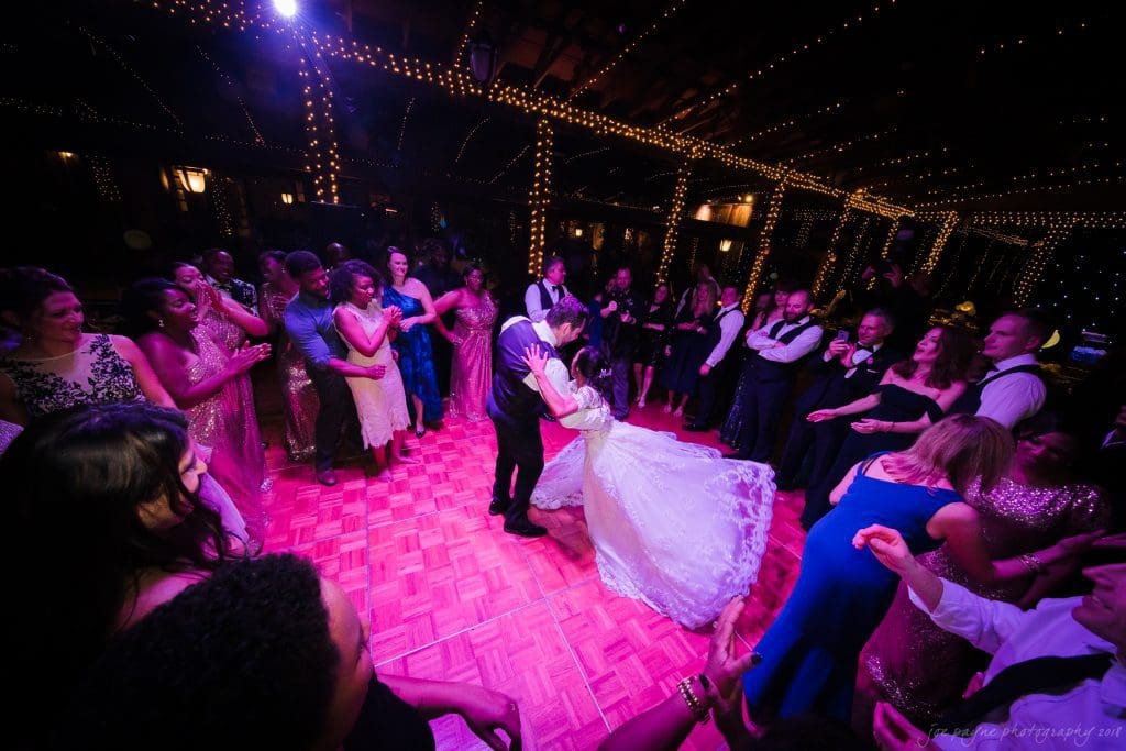 biltmore wedding photographer - anneké & andrew A bride and groom share a romantic dance in the center of a wooden dance floor, surrounded by guests clapping and smiling. The venue is decorated with string lights, creating a warm, festive atmosphere.