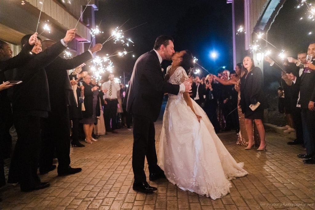 biltmore wedding photographer - anneké & andrew A newlywed couple shares a kiss under a canopy of sparklers held by cheering guests. The bride is in a white gown and the groom in a black suit. The scene is warmly lit with a joyful ambiance.