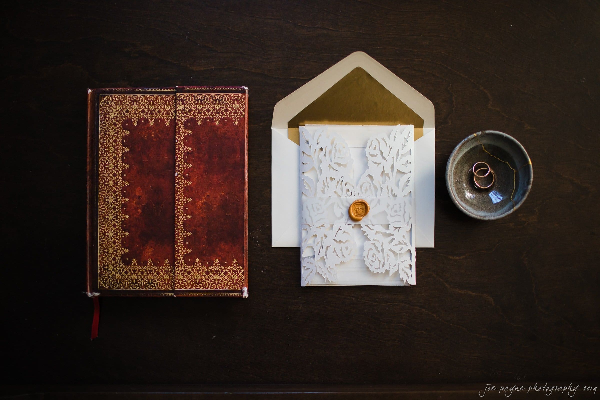 carolina inn & duke chapel wedding - kathleen & michael An ornate red book, a gold-lined envelope with a decorative white card sealed with wax, and two rings in a small dish on a dark wooden surface.