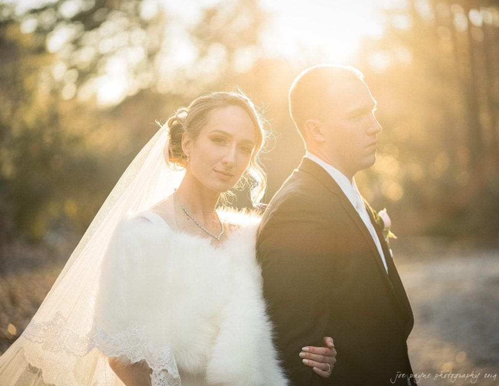 Bride and groom stand together in an outdoor setting illuminated by golden sunlight. The bride wears a veil and fur wrap, while the groom is in a suit. They have a serene, contemplative expression. The background is softly blurred with trees.