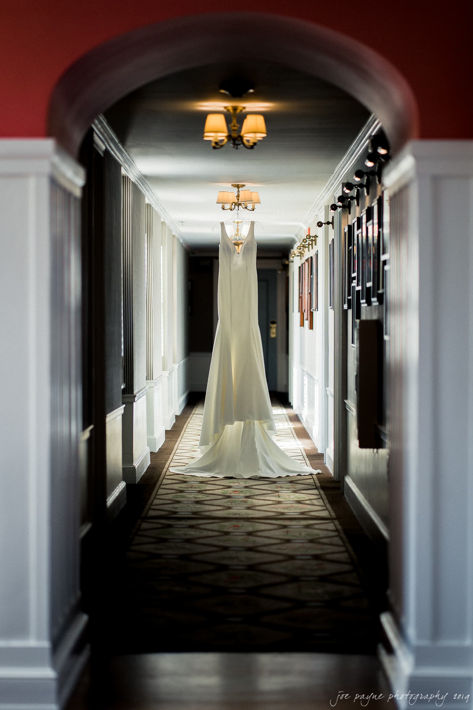carolina inn & duke chapel wedding - kathleen & michael A white wedding dress hangs at the end of a long, narrow hallway with patterned carpet and framed pictures on the walls. The hall is lit by soft light from chandeliers above.