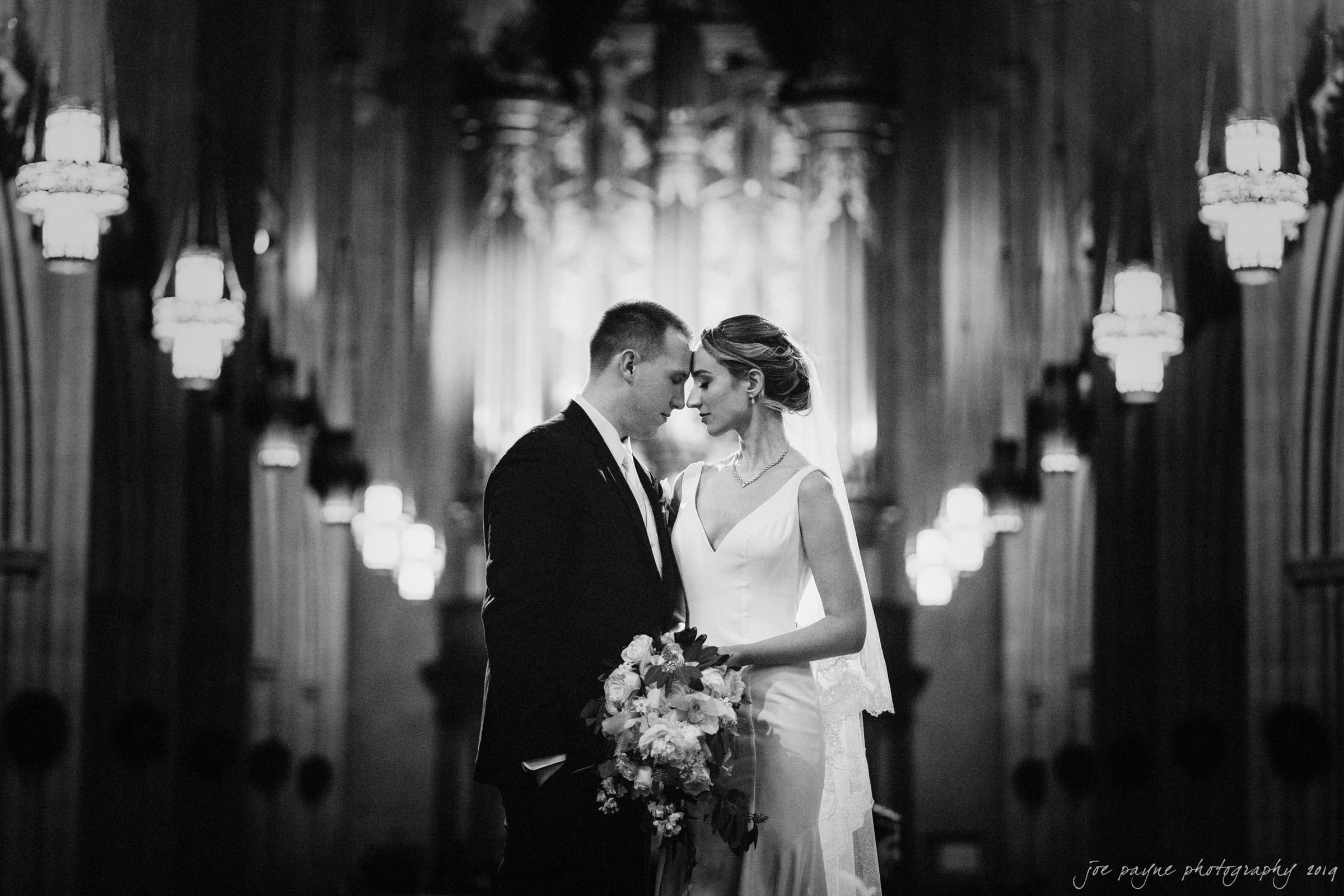 carolina inn & duke chapel wedding - kathleen & michael A bride and groom stand close in an ornate church, illuminated by hanging chandeliers. The bride holds a bouquet of flowers. They share an intimate moment, facing each other, with blurred architectural details in the background. Black and white image.