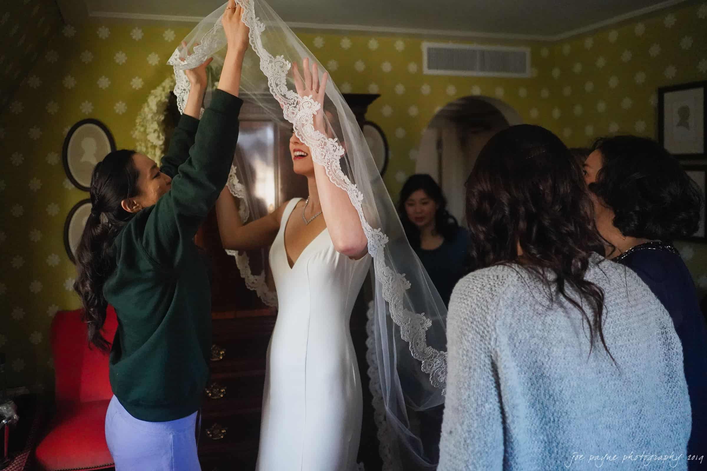 carolina inn & duke chapel wedding - kathleen & michael A bride in a white gown, surrounded by a group of women, adjusts a lace veil over her head. The room is warmly lit with patterned walls and framed portraits. The womens expressions convey joy and excitement.