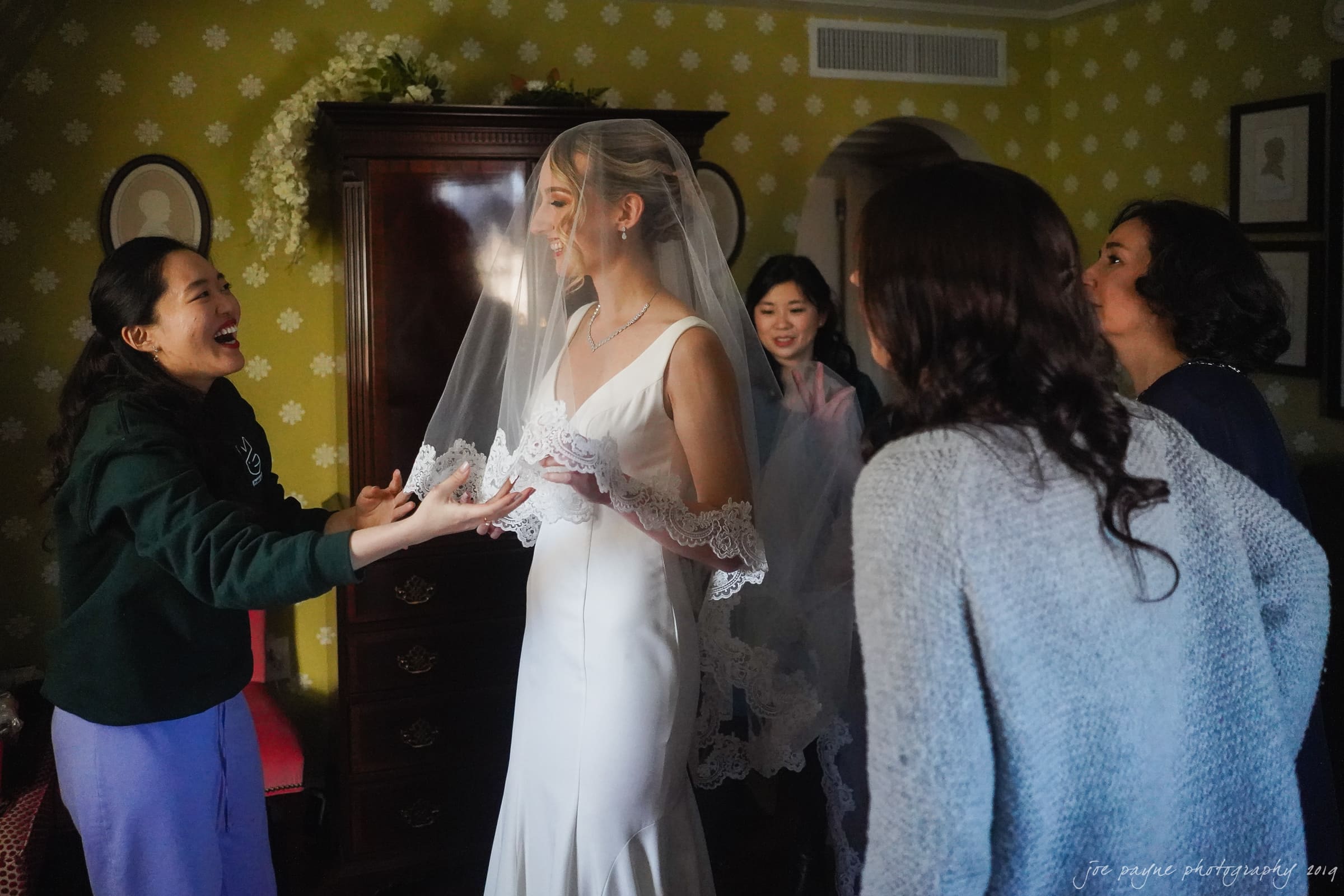 carolina inn & duke chapel wedding - kathleen & michael A bride in a white dress and veil smiles as she stands in a room surrounded by four women. The women appear to be joyfully preparing her for the wedding day, with warm yellow wallpaper in the background.