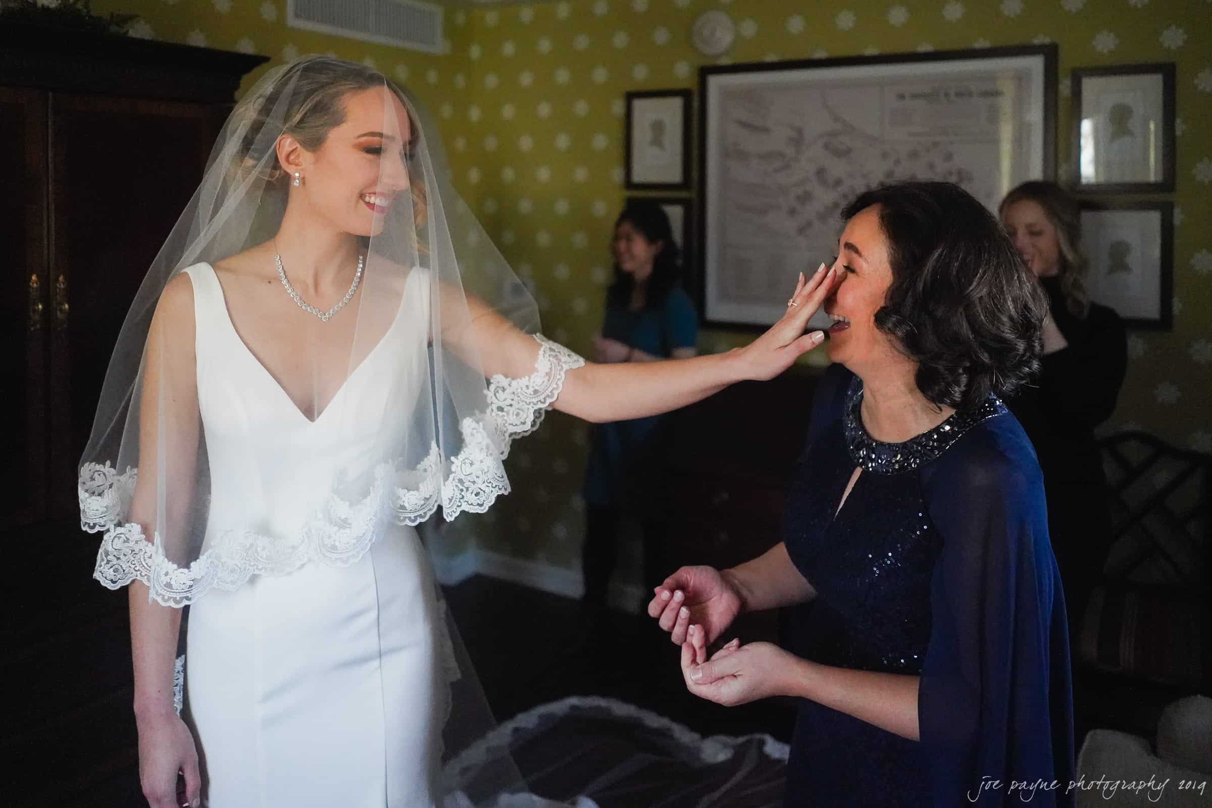 carolina inn & duke chapel wedding - kathleen & michael A bride in a white dress and veil gently touches the face of a woman in blue, both smiling. They stand in a room with green patterned wallpaper, and two other people are in the background.