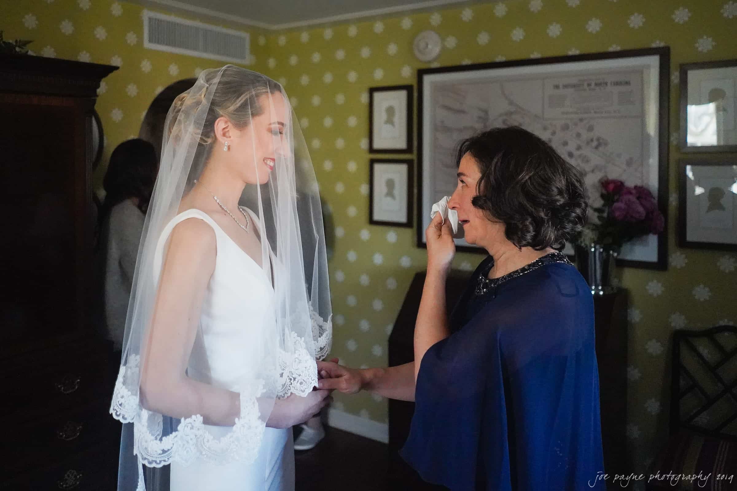 carolina inn & duke chapel wedding - kathleen & michael A bride in a white dress and veil stands with a smiling woman in a dark blue dress, who is wiping tears with a tissue. They are indoors, with framed pictures on the wall and flowers in the background.
