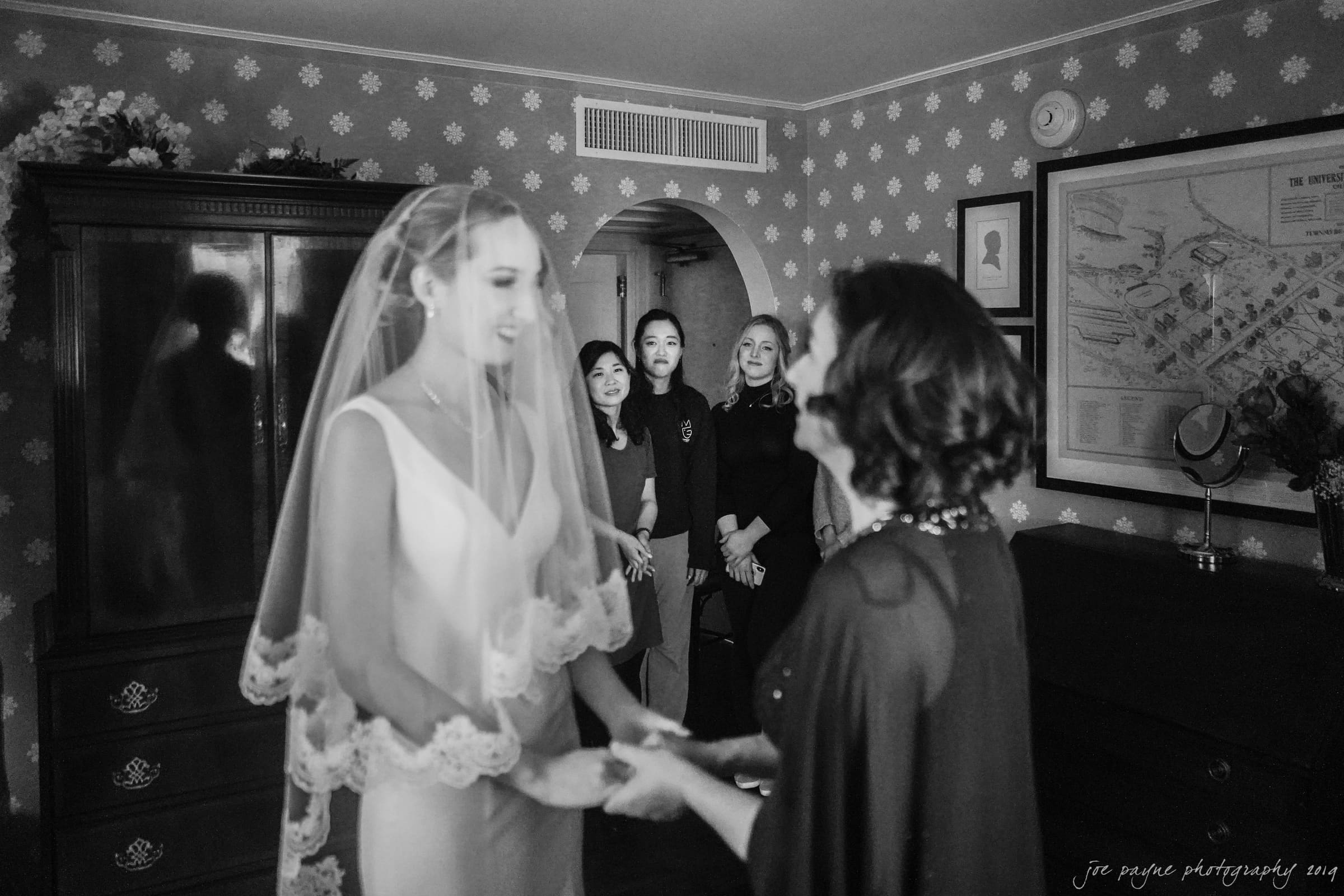 carolina inn & duke chapel wedding - kathleen & michael A bride in a wedding dress and veil smiles while holding hands with another woman. Three women stand behind them in a room with floral wallpaper and vintage decor.