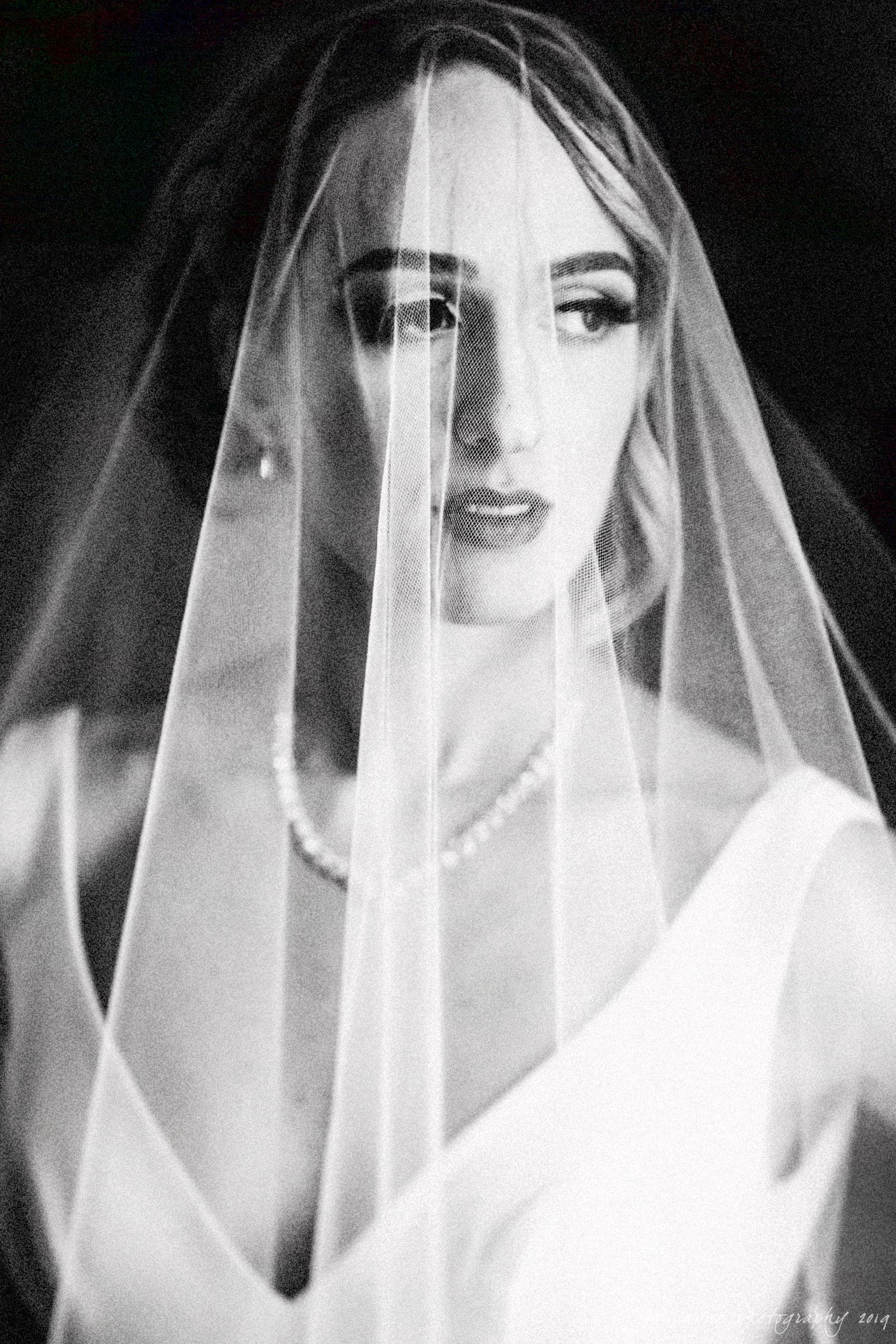 carolina inn & duke chapel wedding - kathleen & michael A black and white portrait of a bride wearing a veil. She is looking to the side, illuminated softly. Her expression is serene, and she is dressed in a white gown with a delicate necklace.