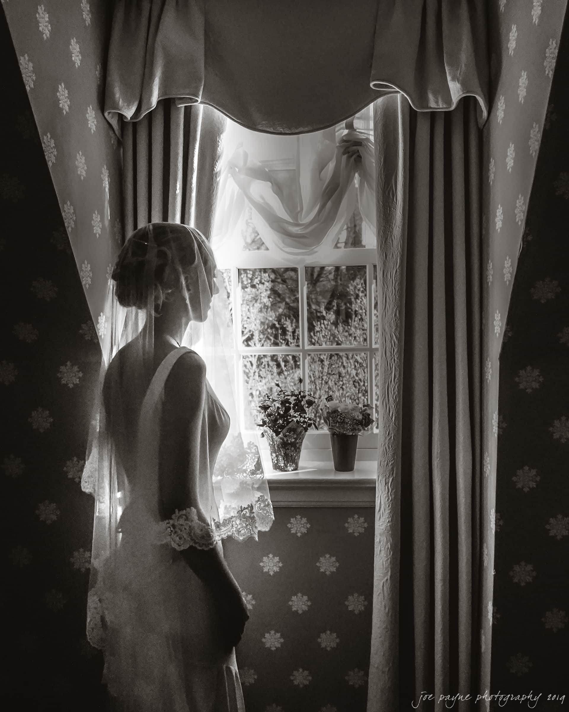 carolina inn & duke chapel wedding - kathleen & michael A bride stands facing a window with sheer curtains, bathed in soft light. She is wearing a delicate veil and a gown with lace details, captured in an elegant, monochromatic style. Floral wallpaper and potted plants create a serene atmosphere.