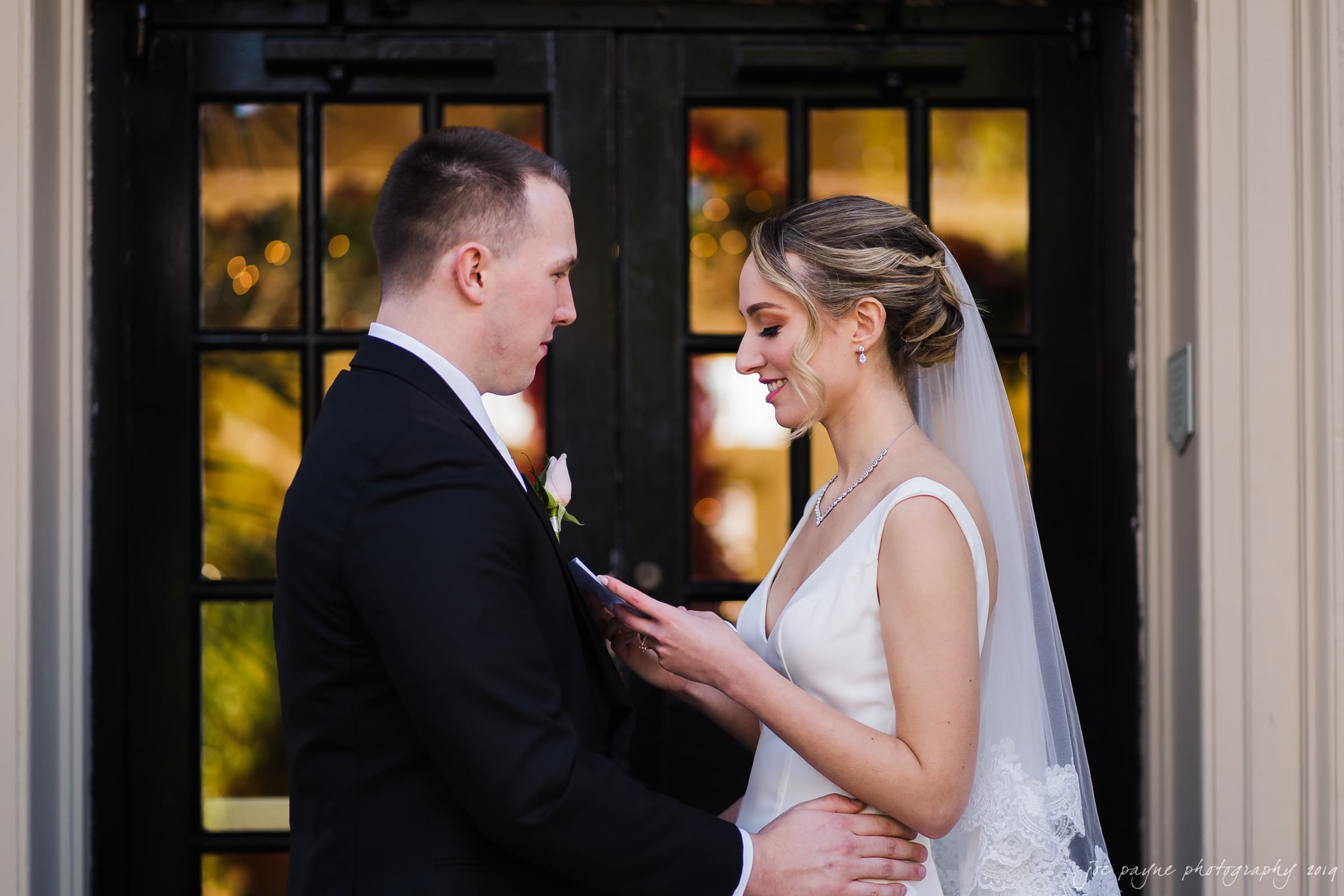 carolina inn & duke chapel wedding - kathleen & michael A bride and groom stand facing each other in front of a black door. The bride, in a white dress and veil, is reading from a paper, while the groom, in a black suit, listens attentively with his hand on her waist.