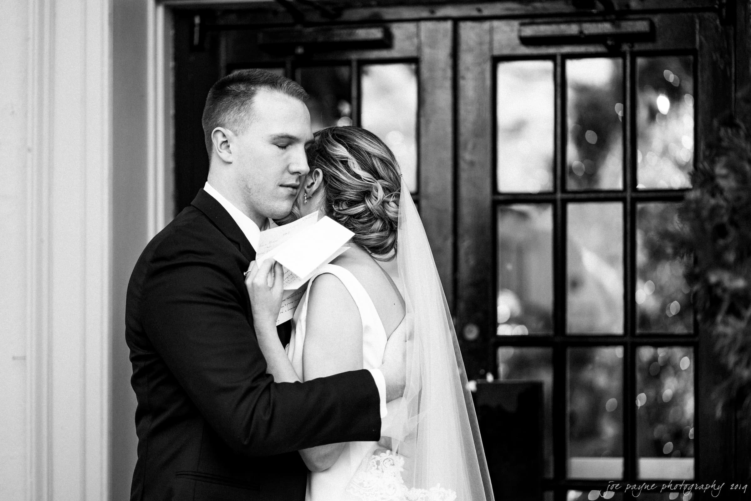 carolina inn & duke chapel wedding - kathleen & michael A black and white photo of a bride and groom embracing tenderly in front of a glass-paneled door. The bride, in a veil, holds a letter, and the groom gently holds her. Both appear emotional in this intimate moment.