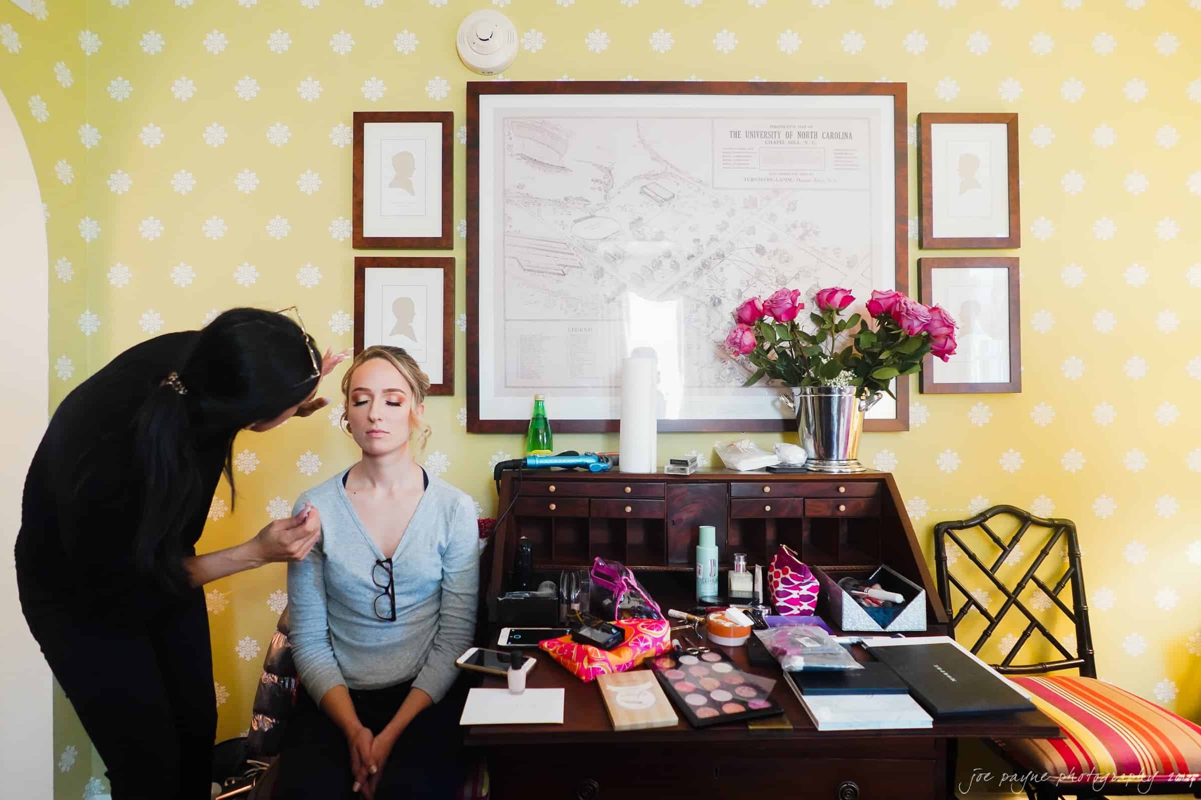 carolina inn & duke chapel wedding - kathleen & michael A woman is having makeup applied by another person in a room with yellow patterned walls. A framed map and portraits hang on the wall. A wooden desk is covered with makeup products and pink roses in a vase.