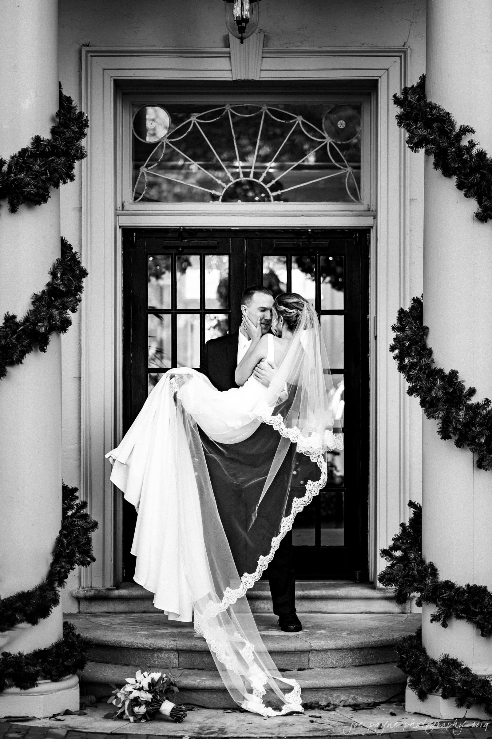 carolina inn & duke chapel wedding - kathleen & michael A groom, in a dark suit, is joyfully lifting a bride, in an elegant white gown and veil, as they kiss in front of a decorated doorway. Greenery adorns the pillars on either side of the entrance. A bouquet rests on the steps.