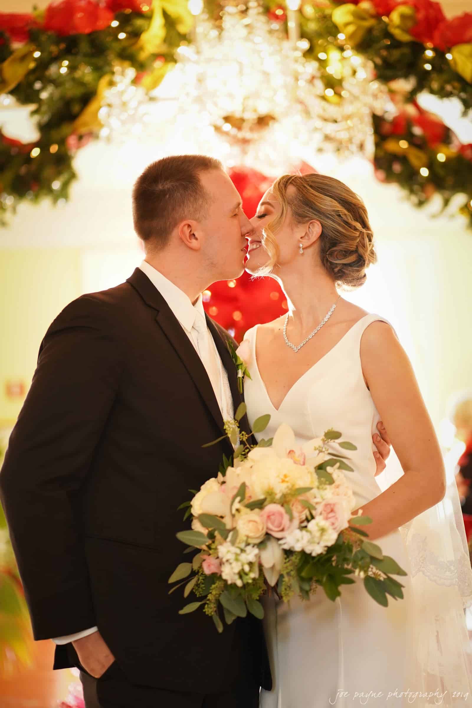 carolina inn & duke chapel wedding - kathleen & michael A bride and groom share a kiss under a decorated chandelier. The bride holds a bouquet of flowers and wears a white gown, while the groom wears a black suit. The background is adorned with festive garlands and lights.