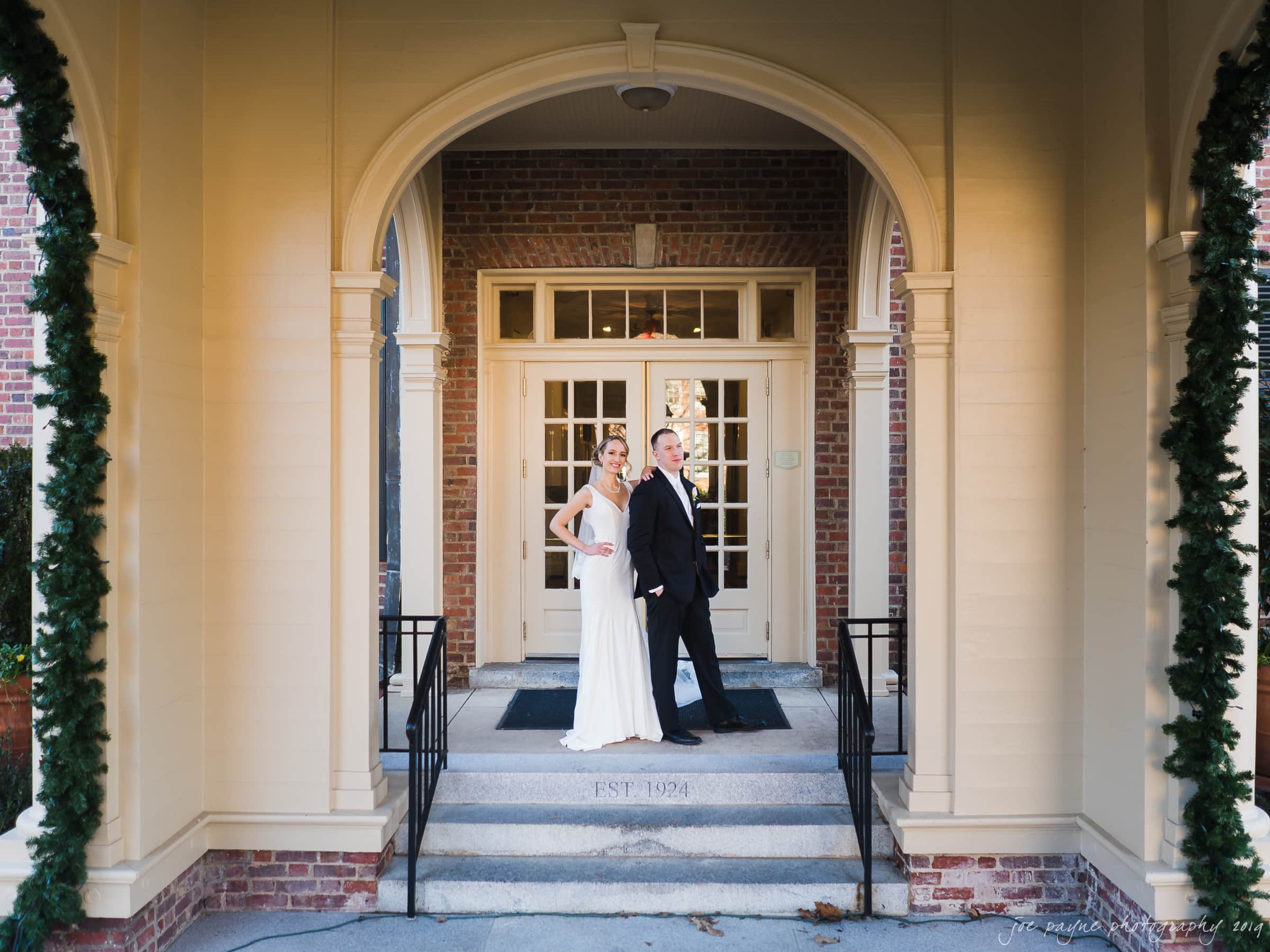 carolina inn & duke chapel wedding - kathleen & michael A couple in formal attire pose on a brick buildings steps. The woman wears a white gown and the man a black suit. The entrance, framed by arches and pillars, features greenery and EST. 1926 on the step.