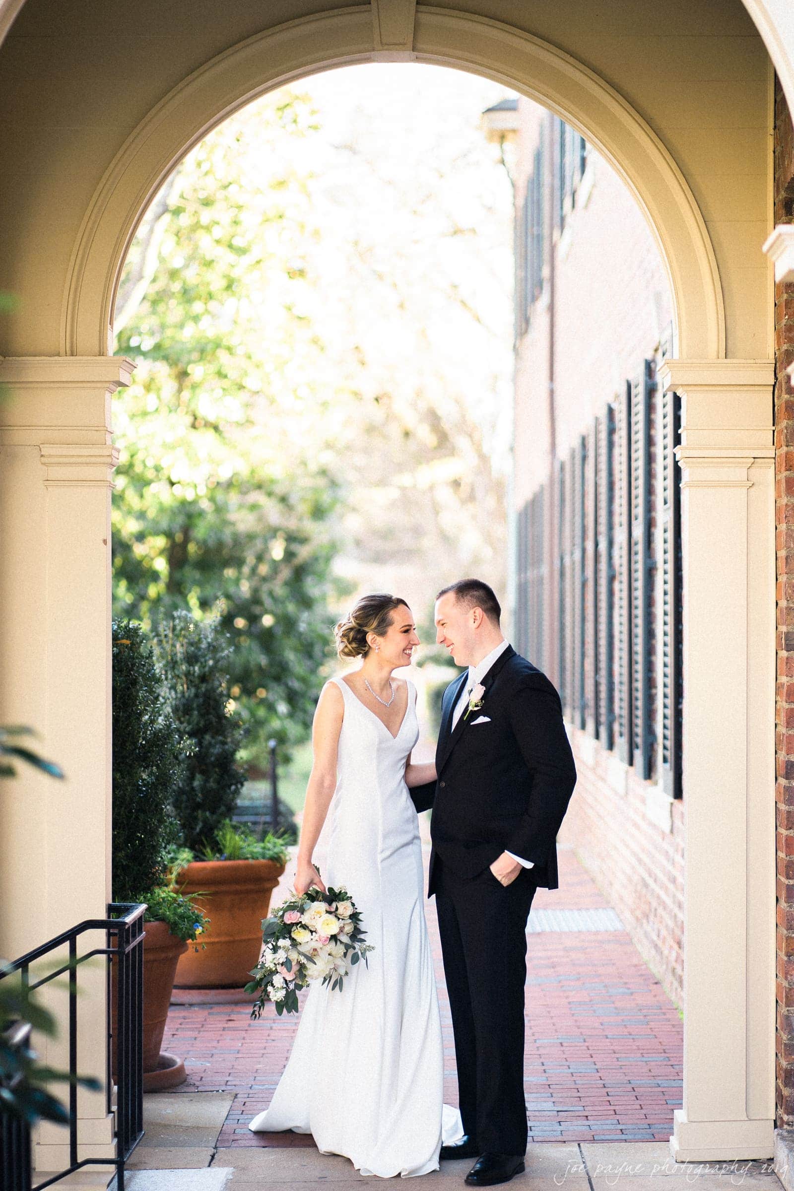 carolina inn & duke chapel wedding - kathleen & michael Bride and groom stand together under an archway, holding a bouquet. She wears a white dress and he a black suit. Sunlight illuminates the brick path and greenery behind them, creating a romantic setting.