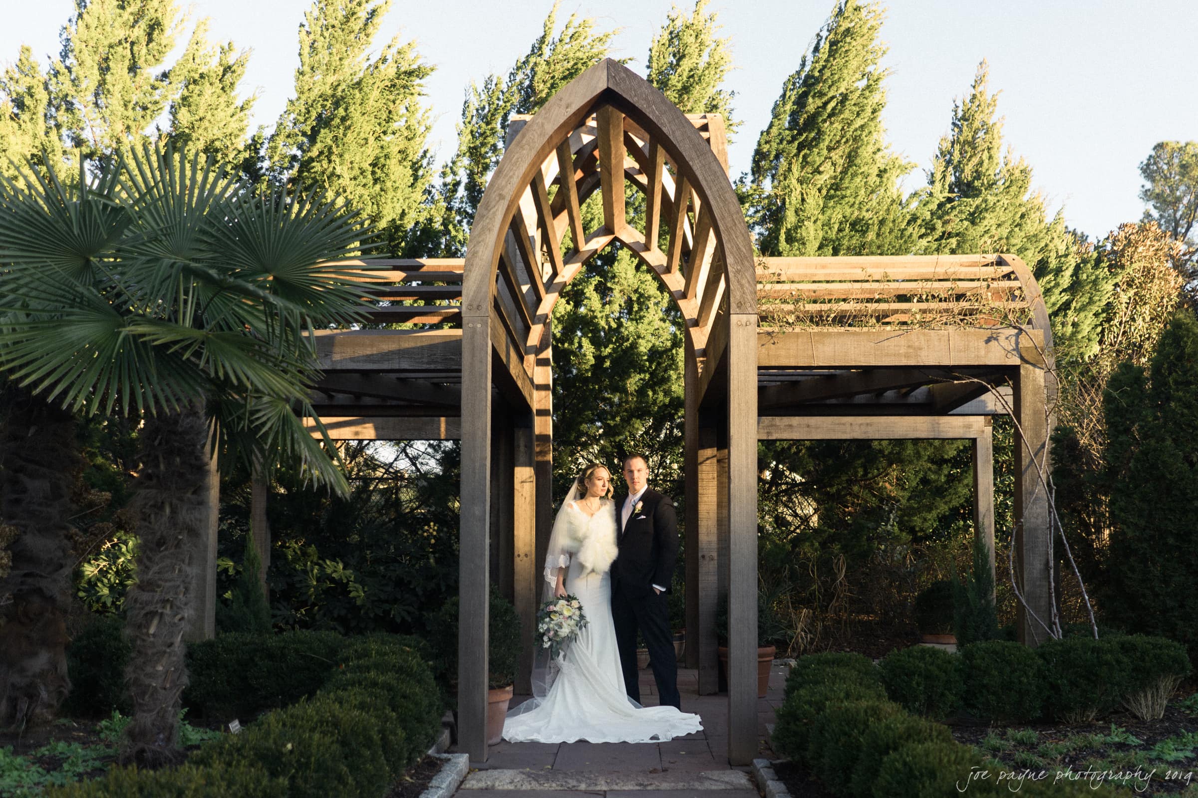 carolina inn & duke chapel wedding - kathleen & michael A bride in a white wedding dress and a groom in a black suit stand together under a wooden archway in a lush garden setting, surrounded by greenery and trees.