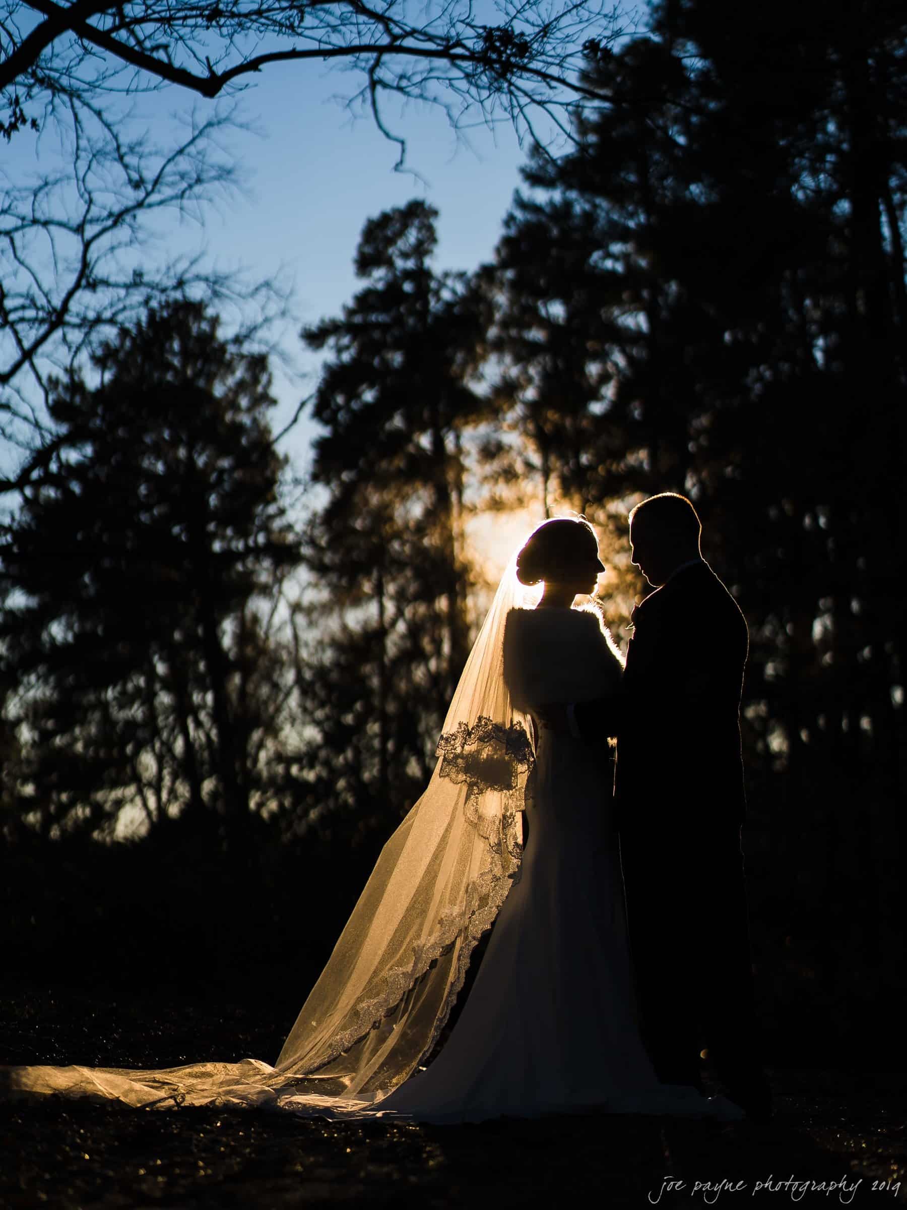 carolina inn & duke chapel wedding - kathleen & michael Silhouette of a couple in wedding attire standing close together outdoors, with the sun setting behind them and trees in the background. The brides veil is flowing and highlighted by the sun.