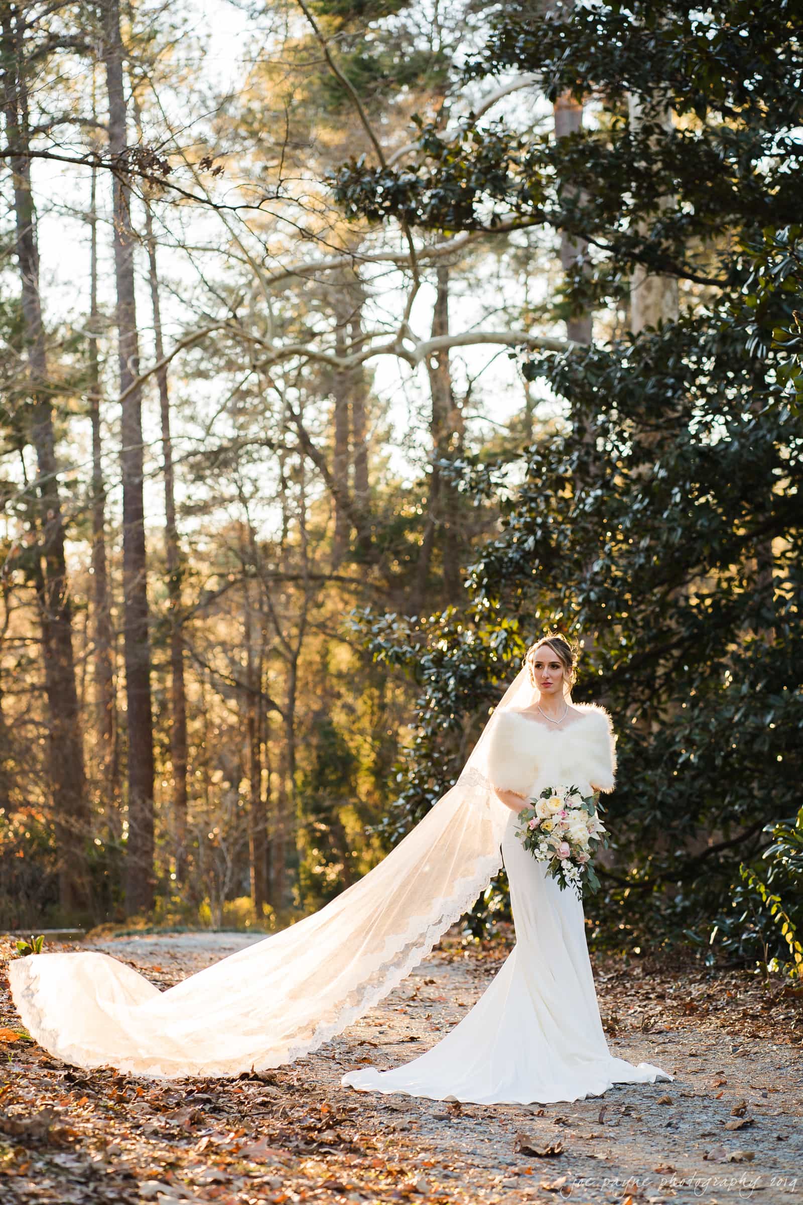 carolina inn & duke chapel wedding - kathleen & michael A bride stands on a sunlit path in a forest, wearing a long white gown with an extended lace train. She holds a bouquet of flowers and the sun filters through the trees, creating a serene and romantic atmosphere.