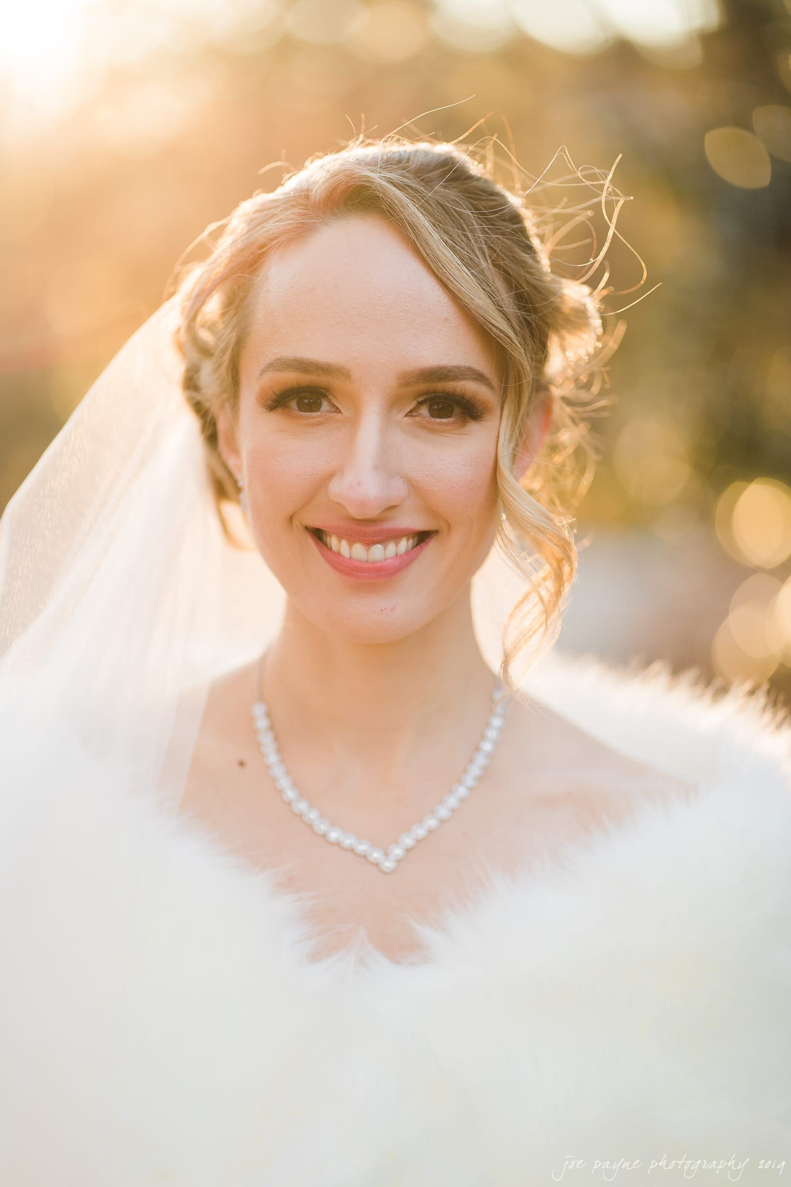 carolina inn & duke chapel wedding - kathleen & michael A bride wearing a pearl necklace and a veil smiles warmly. The background features soft, golden bokeh from sunlight filtering through trees, creating a dreamy and romantic atmosphere.