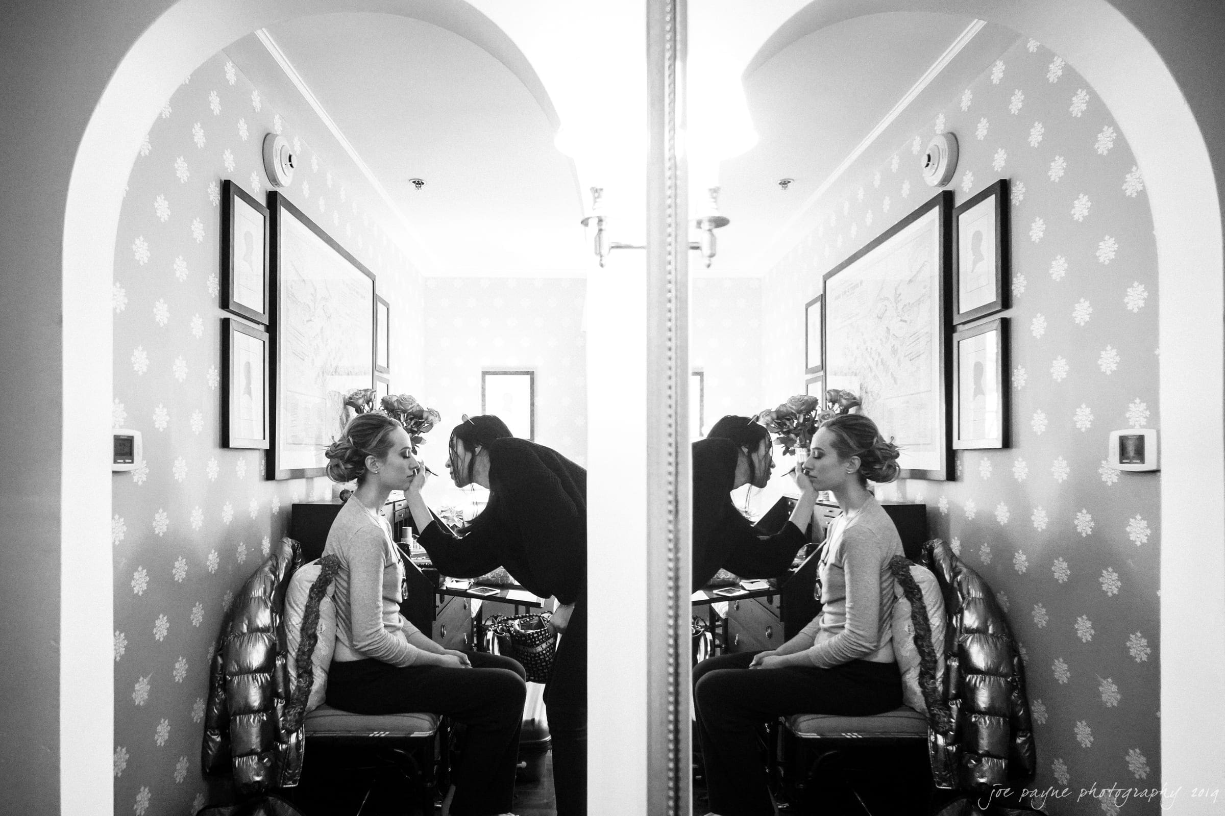 carolina inn & duke chapel wedding - kathleen & michael A black and white image of a woman sitting and getting her makeup done by another person. They are seated in front of a mirror, creating a symmetrical reflection. The room is decorated with patterned wallpaper and framed artwork.