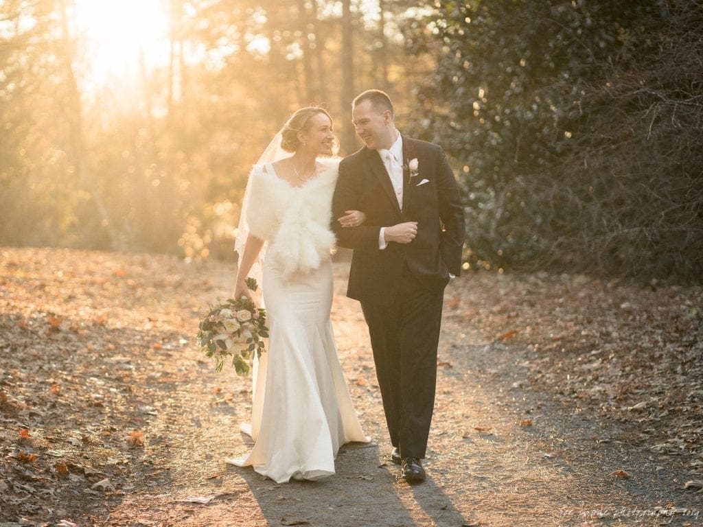 A bride and groom walk arm in arm on a sunlit path. The bride wears a white dress and fur shawl, holding a bouquet. The groom wears a dark suit. Surrounded by autumn leaves, they appear joyful and content.