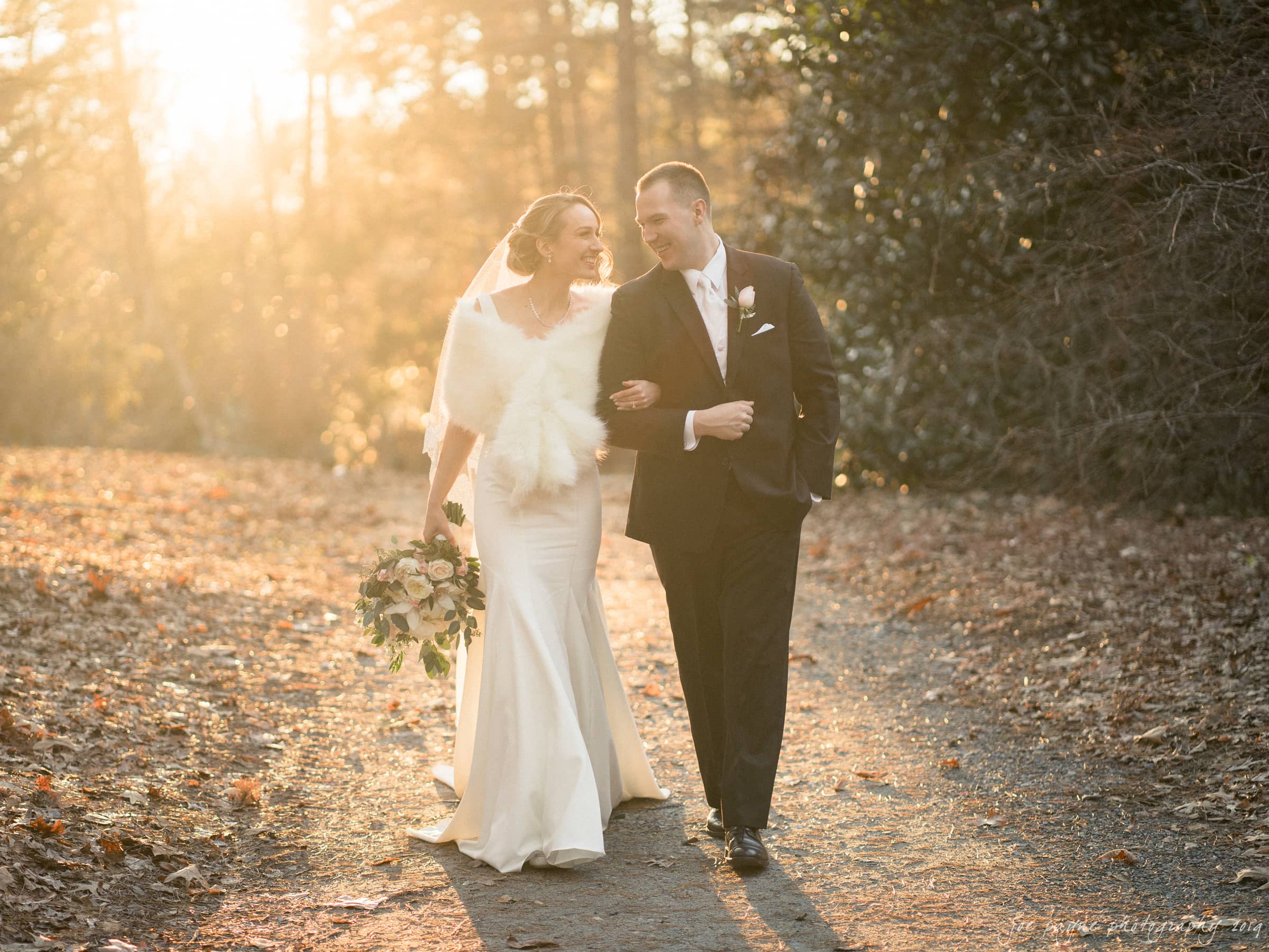 carolina inn & duke chapel wedding - kathleen & michael A bride and groom walk arm in arm on a sunlit path. The bride wears a white dress and fur shawl, holding a bouquet. The groom wears a dark suit. Surrounded by autumn leaves, they appear joyful and content.