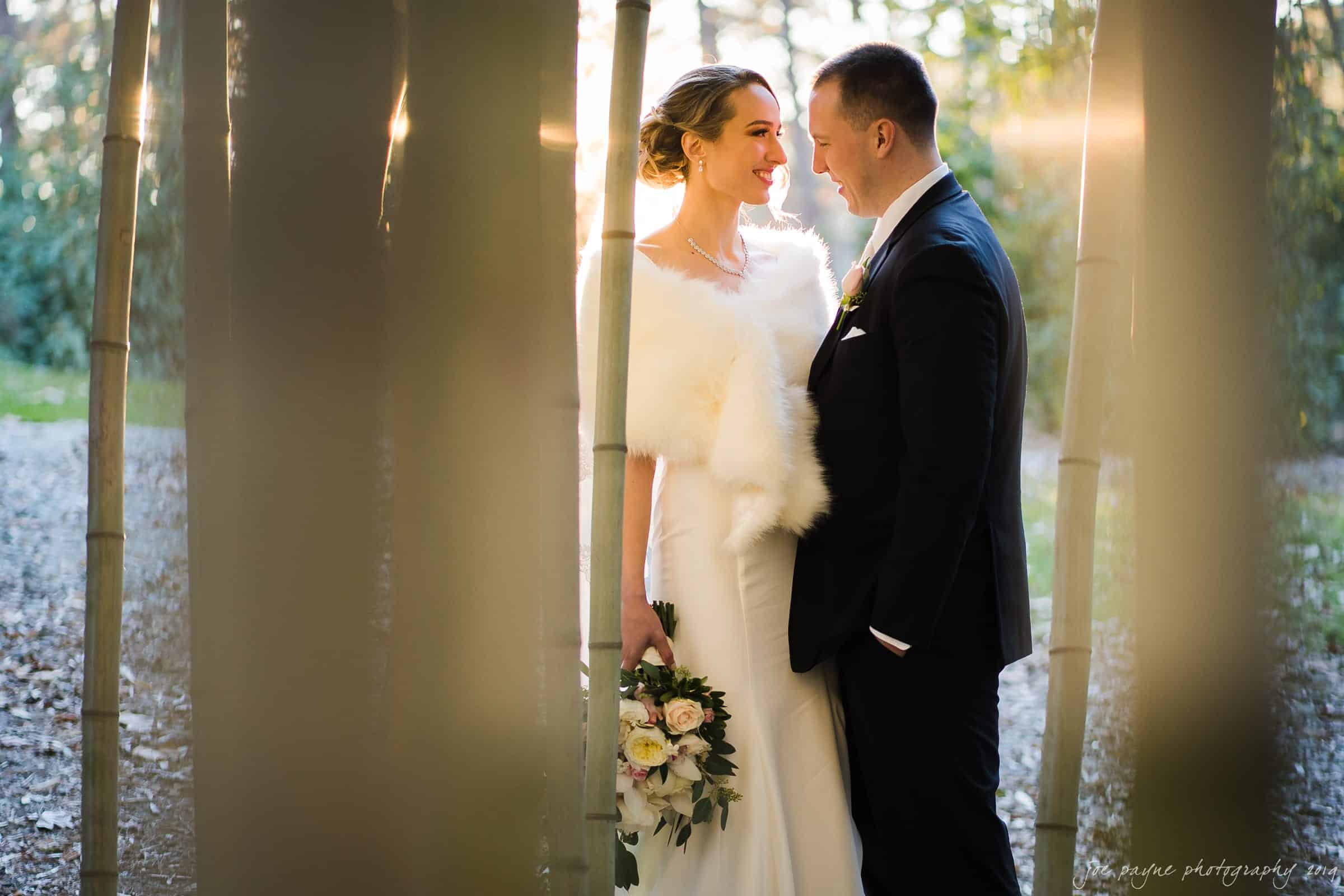 carolina inn & duke chapel wedding - kathleen & michael A bride and groom stand closely together among bamboo stalks. The bride wears a white dress and fur stole, holding a bouquet of flowers, while the groom is in a dark suit with a boutonniere. Soft light filters through the trees behind them.