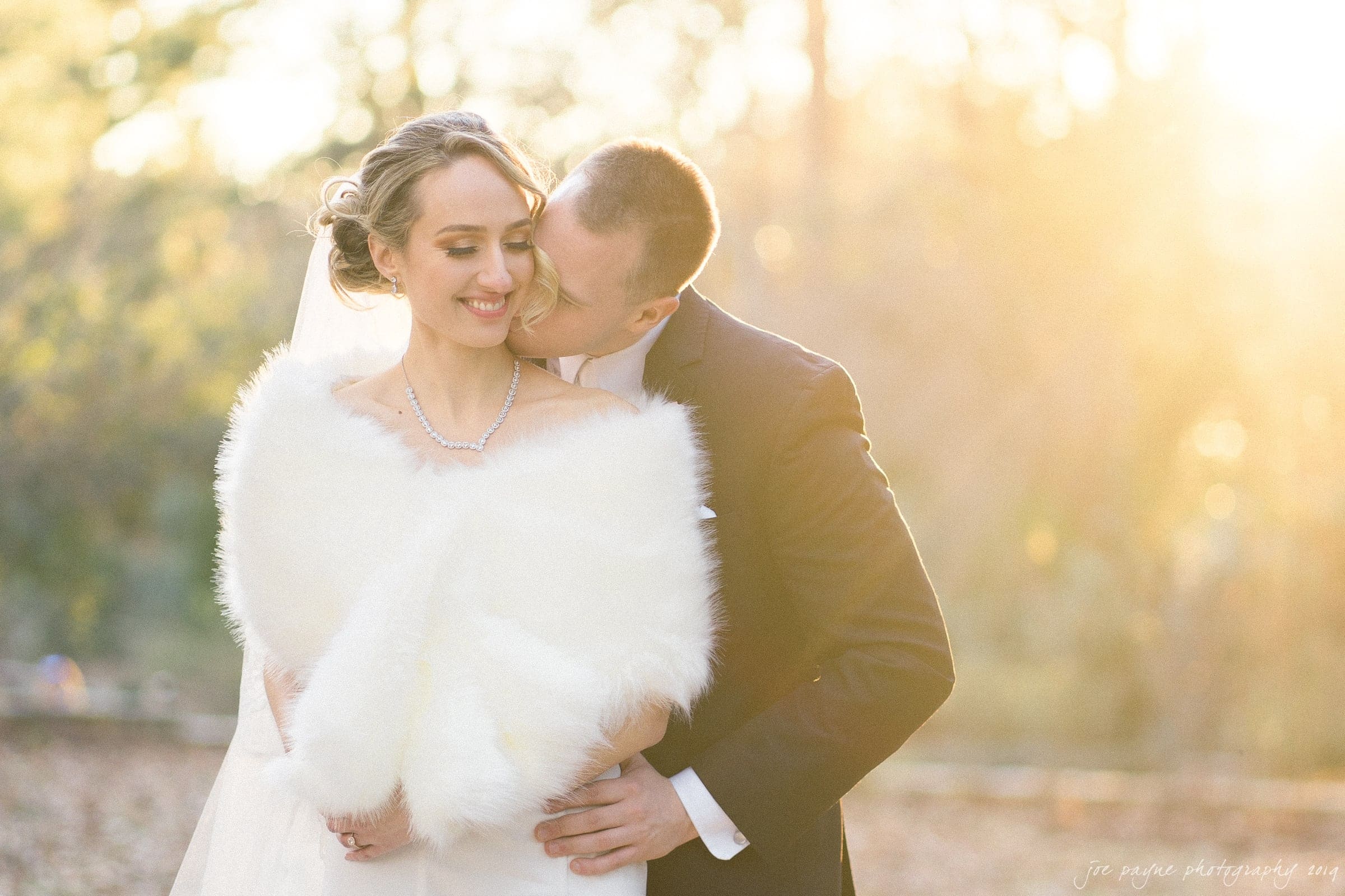 carolina inn & duke chapel wedding - kathleen & michael A couple stands outdoors in soft sunlight, surrounded by trees. The woman is wearing a white fur wrap and veil, smiling, while the man in a suit gently kisses her head. Warm light creates a dreamy atmosphere.