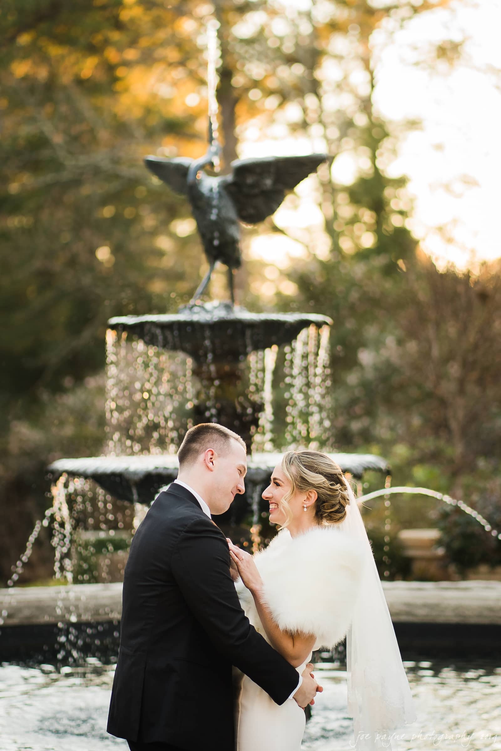 carolina inn & duke chapel wedding - kathleen & michael A couple stands in front of a tiered fountain, sharing a tender moment. The woman is in a white gown with a fur wrap, and the man is in a black suit. Sunlight filters through the trees in the background, creating a warm and romantic atmosphere.