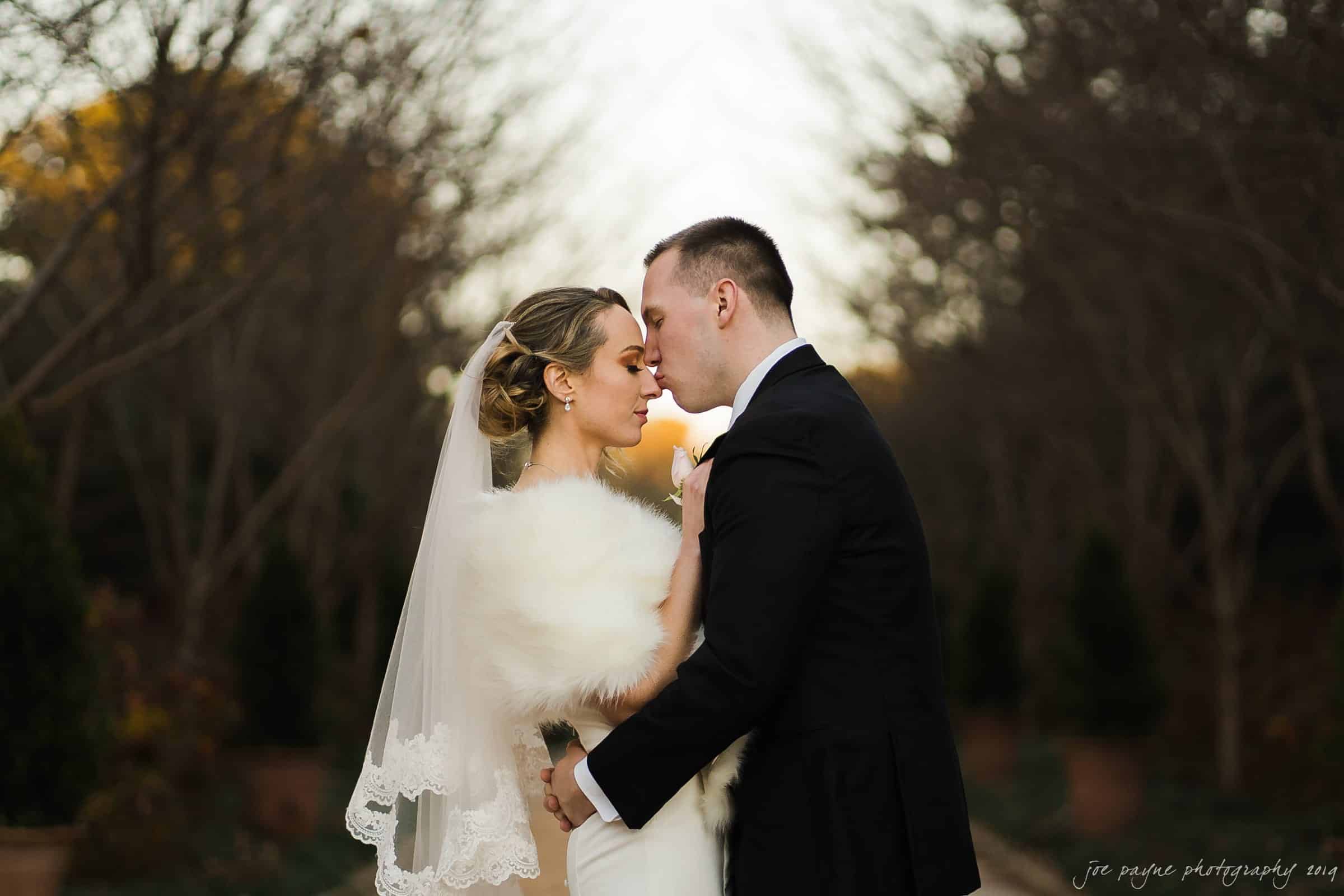 carolina inn & duke chapel wedding - kathleen & michael A bride and groom embrace outdoors. The bride wears a white dress and veil, with a fur shawl. The groom is in a black suit. Bare trees and soft evening light create a romantic atmosphere.