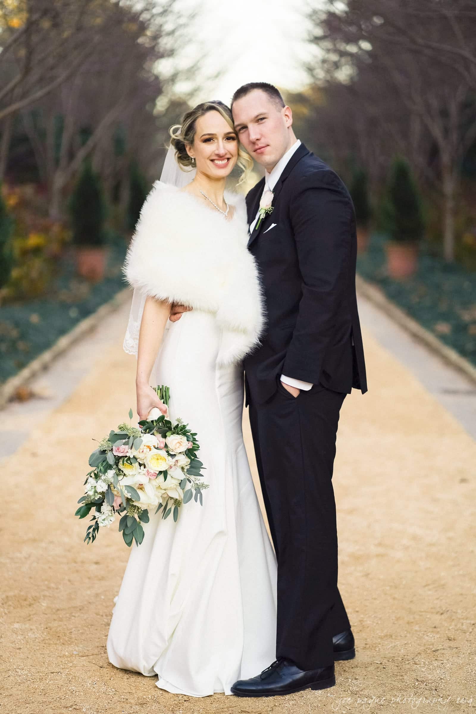 carolina inn & duke chapel wedding - kathleen & michael A bride and groom stand on a tree-lined path. The bride is in a long white dress, holding a bouquet, and wearing a fluffy white shawl. The groom is in a black suit and tie. They both smile at the camera, with greenery visible in the background.