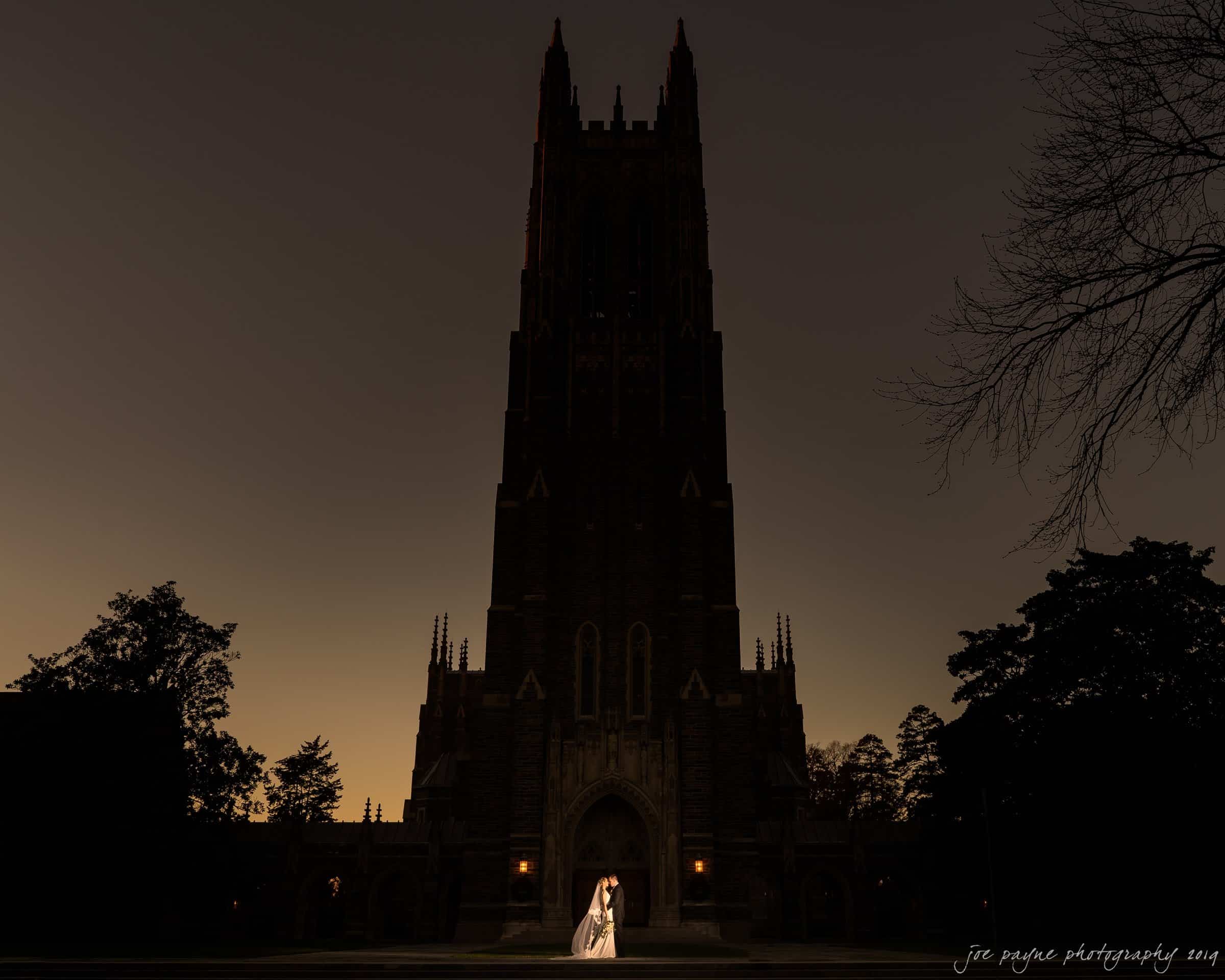 carolina inn & duke chapel wedding - kathleen & michael A couple stands in front of a grand, illuminated cathedral at dusk. The silhouette of the tall building is highlighted against a darkening sky, creating a dramatic and romantic scene.