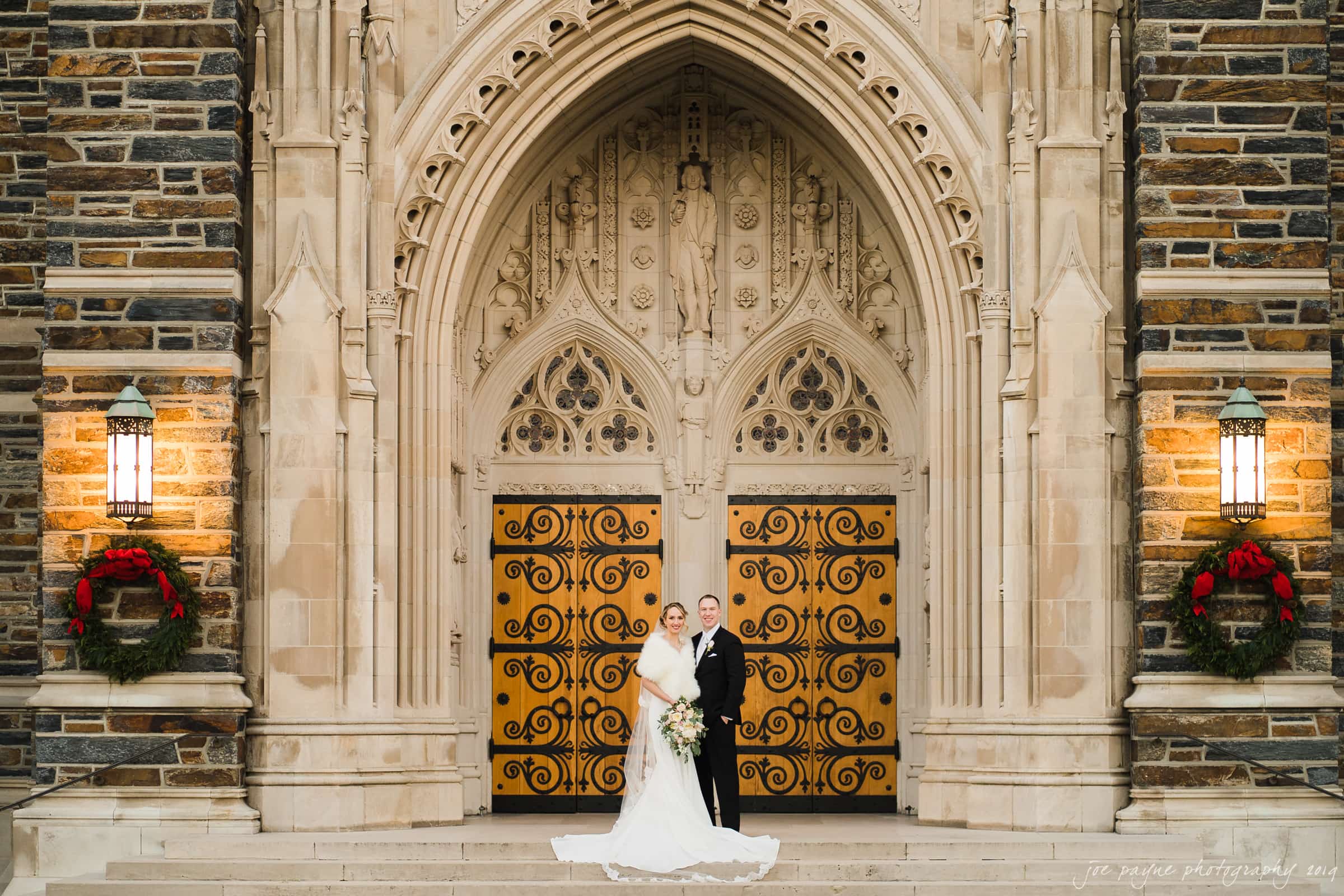 carolina inn & duke chapel wedding - kathleen & michael A bride and groom stand on the steps of a historic stone church with large wooden doors. The bride wears a long white gown, and the groom is in a black suit. Two wreaths adorn the walls, and ornate architecture frames the scene.