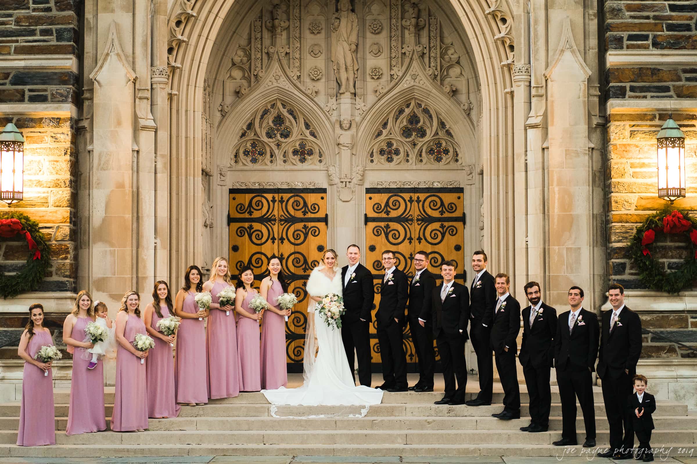 carolina inn & duke chapel wedding - kathleen & michael A wedding party stands in front of a large ornate wooden church door. The bride and groom are in the center, surrounded by bridesmaids in pink dresses and groomsmen in black suits. There are holiday wreaths and lanterns on either side of the door.