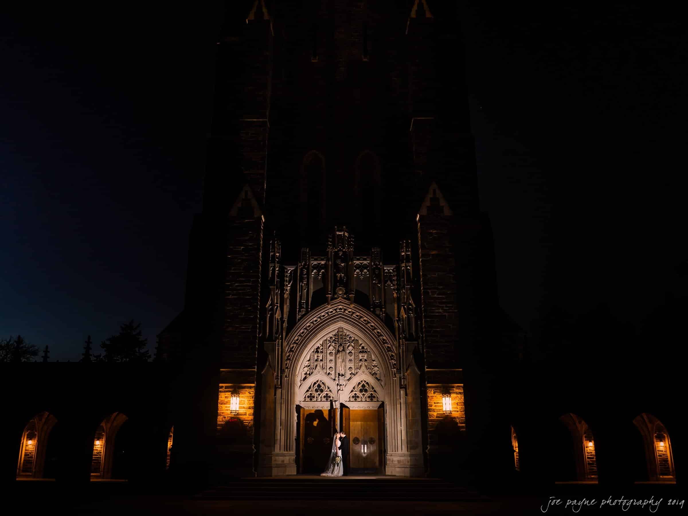 carolina inn & duke chapel wedding - kathleen & michael A couple stands in front of a grand, illuminated church entrance at night. The ornate architecture is highlighted by soft lighting, creating a dramatic contrast with the dark surroundings.