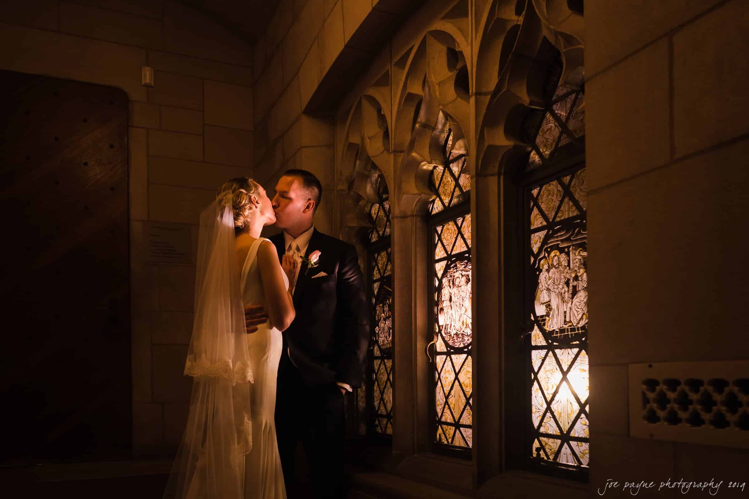 carolina inn & duke chapel wedding - kathleen & michael A bride and groom share an intimate moment in a dimly lit, ornate stone hallway. Stained glass windows illuminate them with a warm glow, highlighting the brides veil and gown as they gaze into each others eyes.