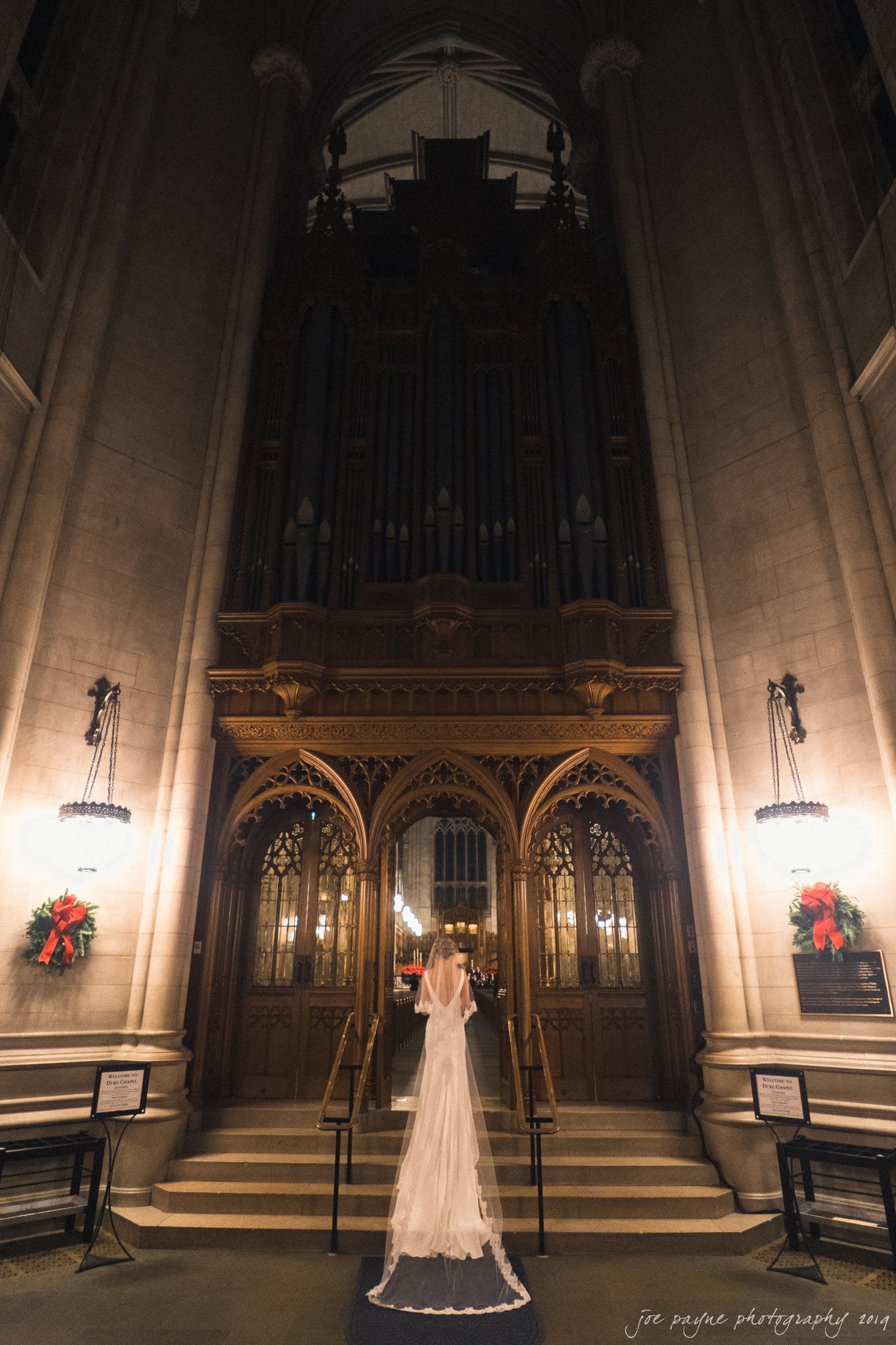 carolina inn & duke chapel wedding - kathleen & michael A bride in a flowing white dress stands before an ornate cathedral entrance, lit by warm ambient lighting. Large wooden doors and an impressive pipe organ are in the background, creating a majestic and serene atmosphere.