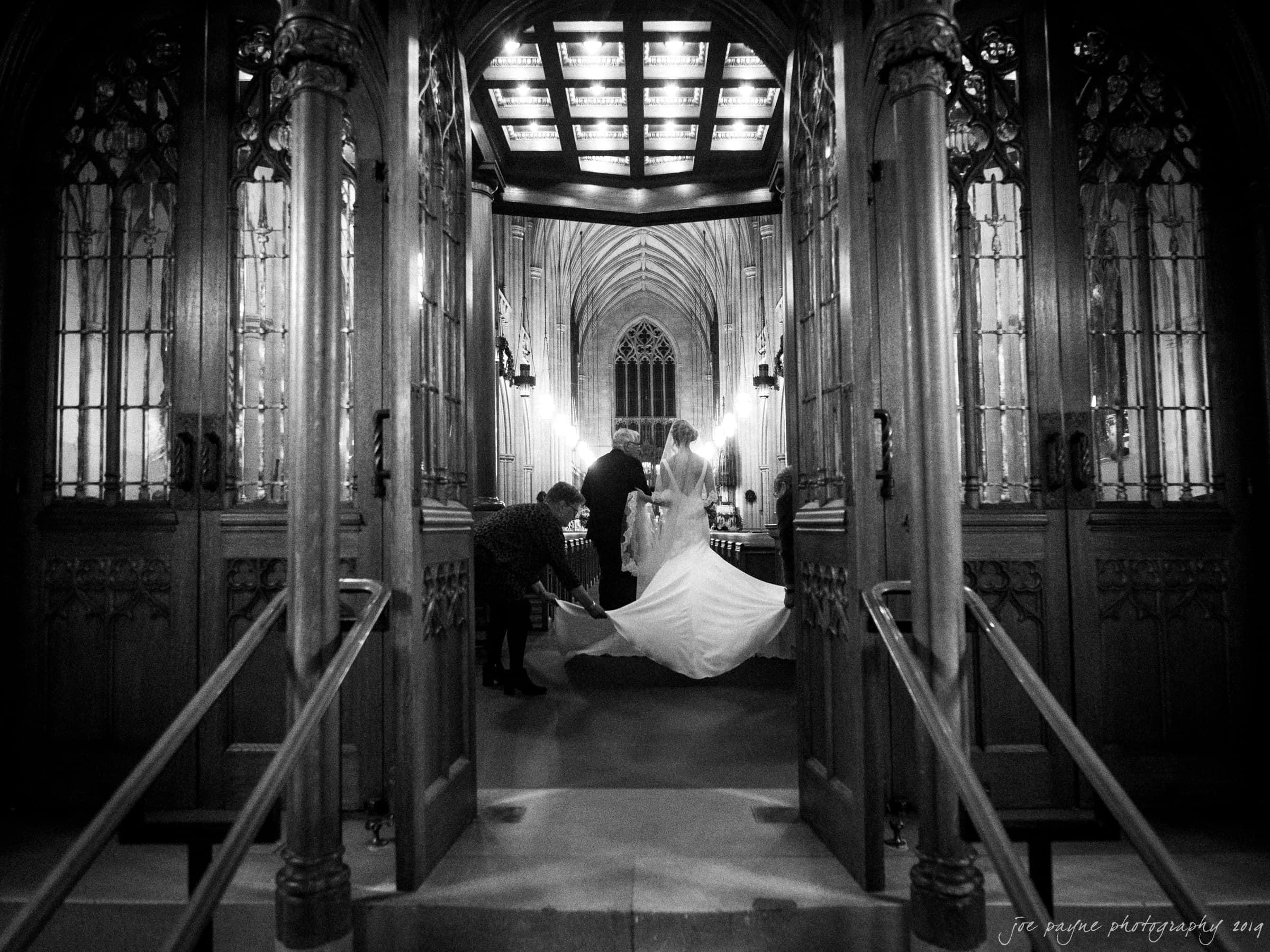 carolina inn & duke chapel wedding - kathleen & michael A black and white photo of a bride and groom standing inside a grand, ornate chapel. The brides dress train is being adjusted by an attendant while light streams down from above, illuminating the couple. Gothic architectural details surround them.