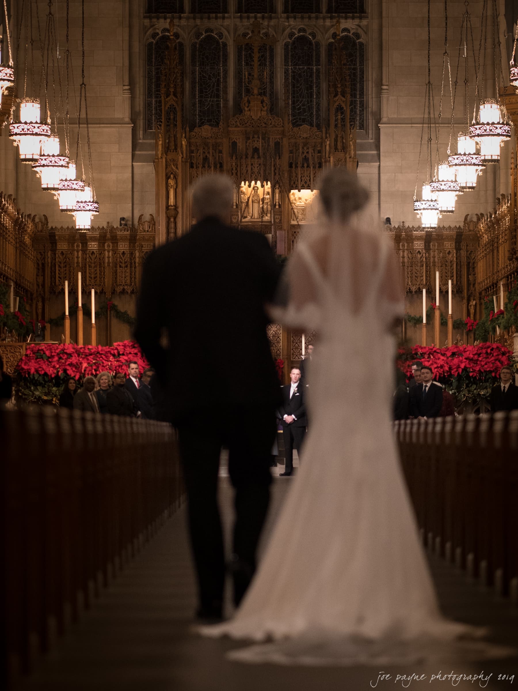 carolina inn & duke chapel wedding - kathleen & michael A bride in a white gown walks down the aisle with an older man in a dark suit inside a grand church. The aisle is lined with red flowers, and decorative lights hang from the ceiling.
