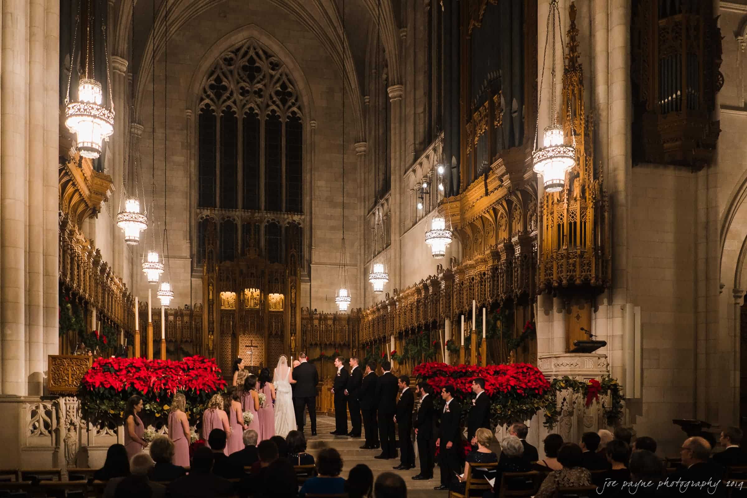carolina inn & duke chapel wedding - kathleen & michael A wedding ceremony inside a grand cathedral with ornate architecture. The bridal party stands near the altar, surrounded by red poinsettias. Guests seated in rows watch the proceedings under the warm glow of hanging chandeliers.